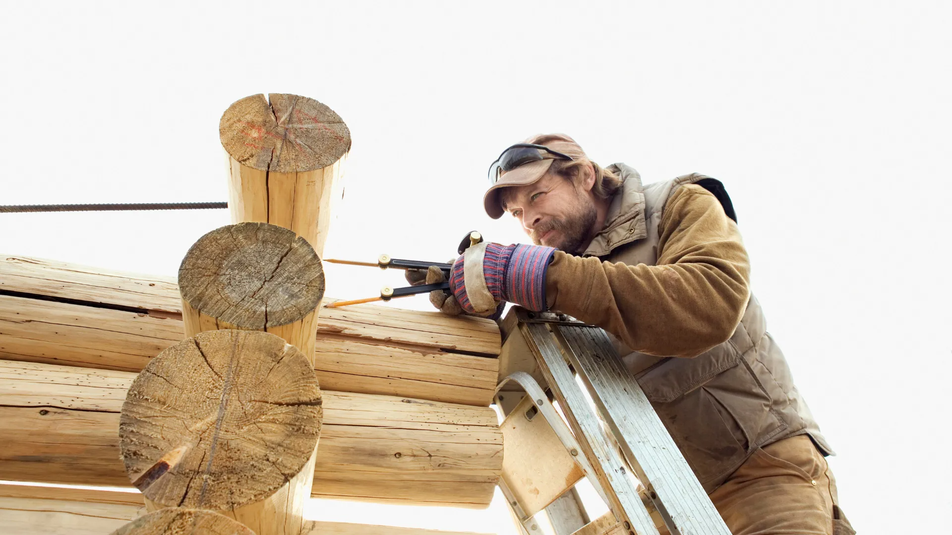 A worker on a ladder uses a marking tool to measure log ends while building a log cabin.