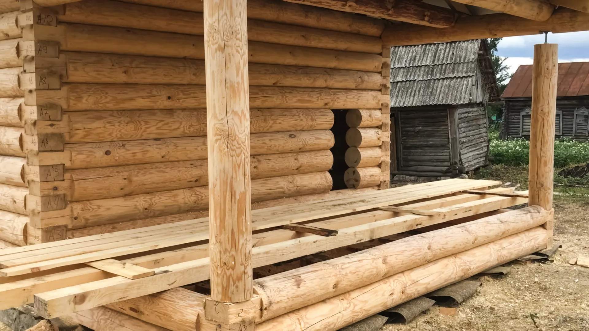 A partially constructed log cabin under a roof, featuring horizontal wall logs and an unfinished wooden porch floor.