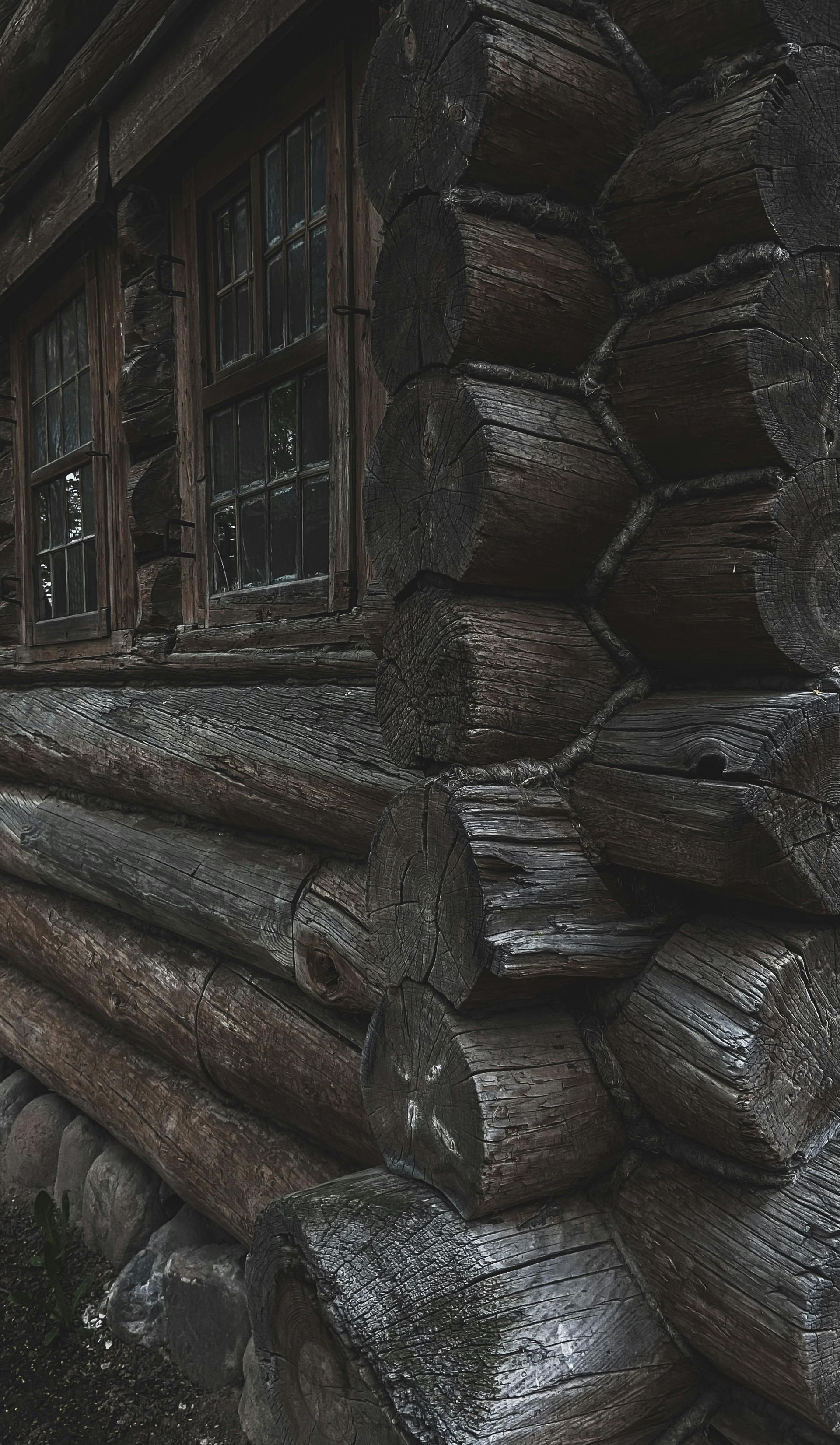 A close-up, angled view of a dark, rustic log cabin wall with visible wood grain, weathered texture, and small windows.