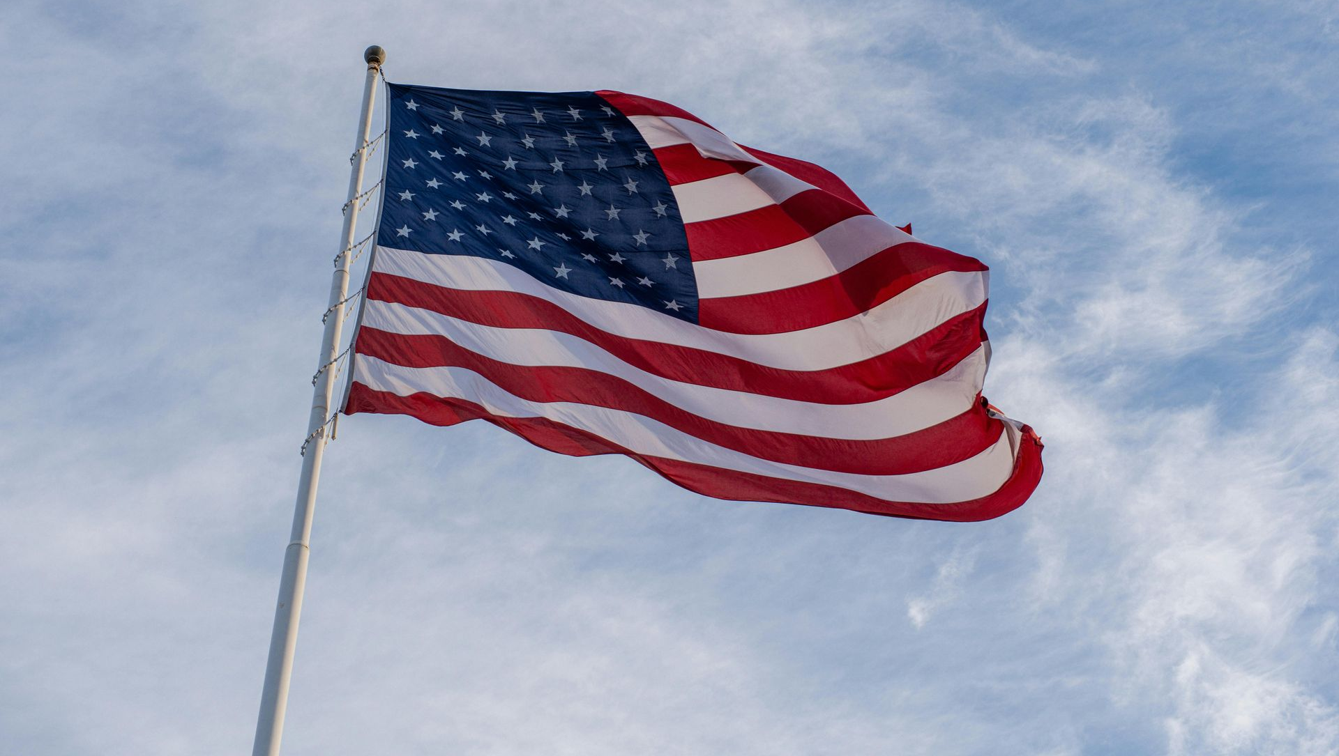 The American flag ripples against a bright, cloudy blue sky.