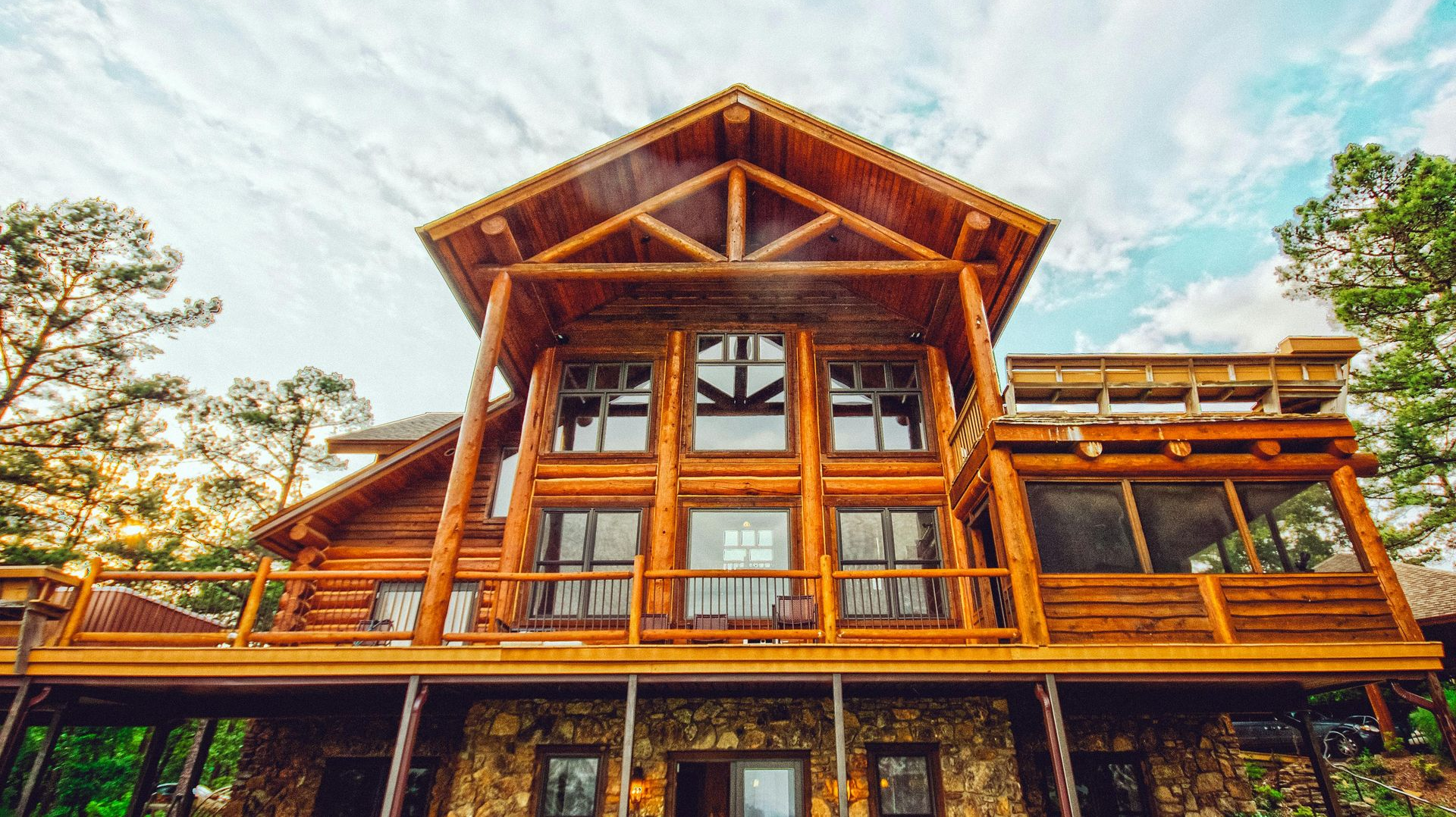 A large, multi-story log cabin with a stone base and wooden balcony, surrounded by trees under a cloudy sky.