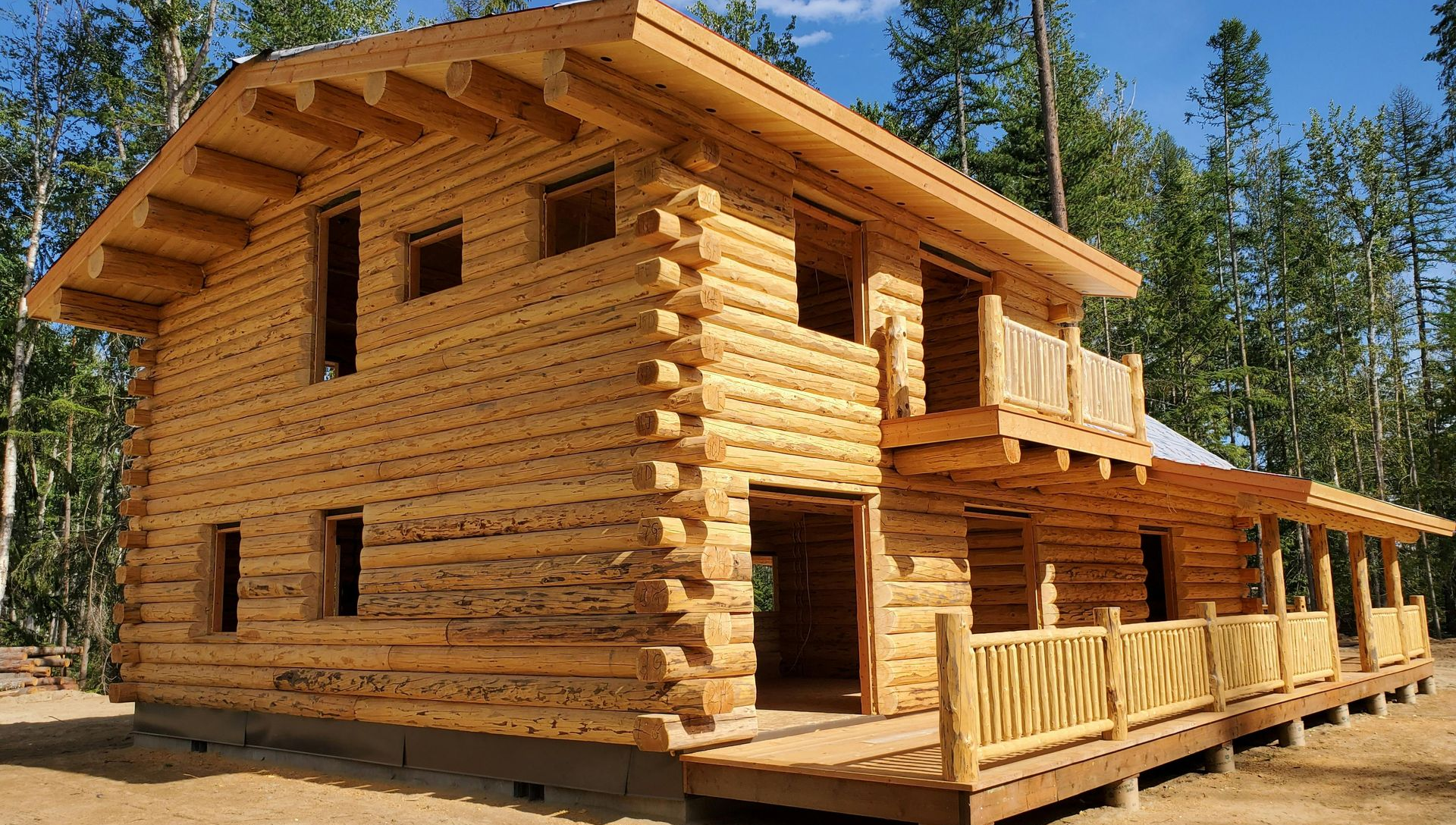 A two-story log cabin under construction with a wraparound porch, surrounded by a forest on a sunny day.