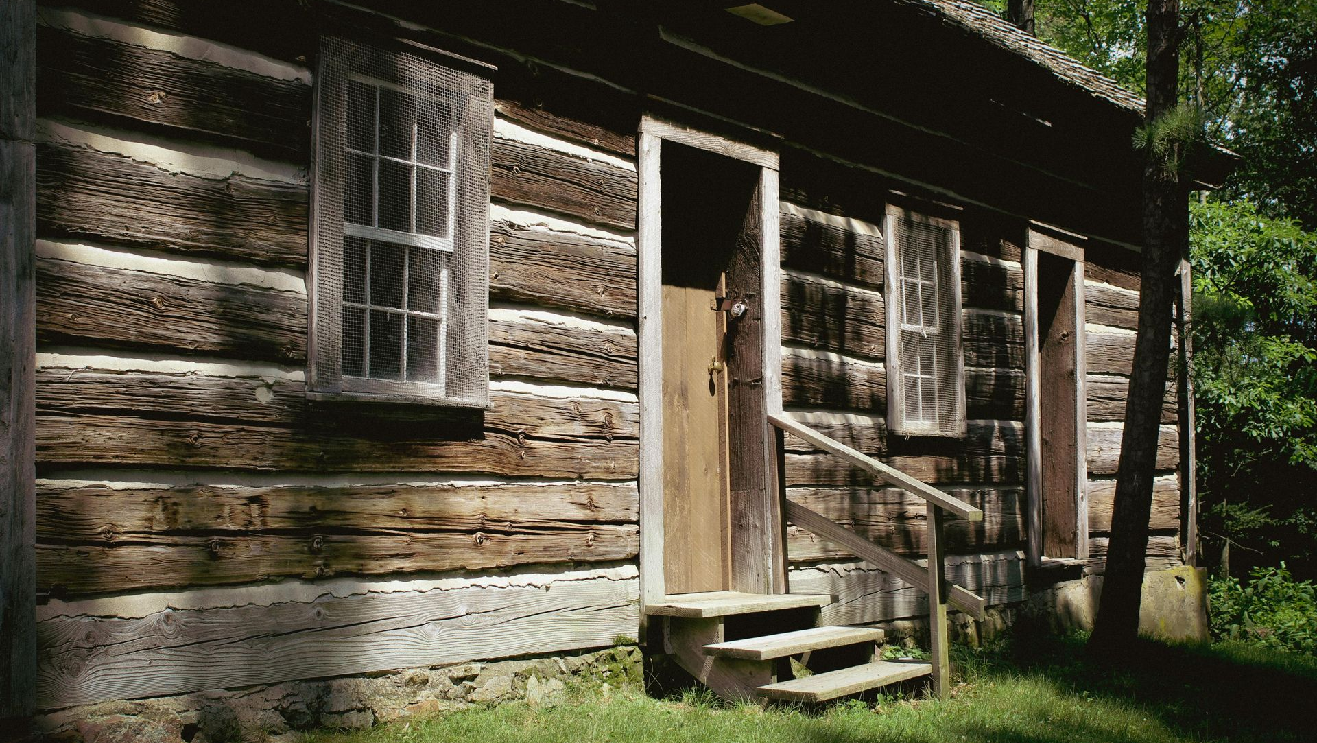 A rustic, dark-wood log cabin with two windows and an open door reached by wooden steps, surrounded by green trees.