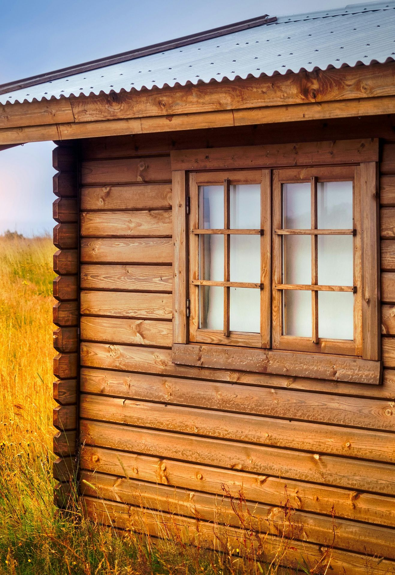 A log cabin wall with a wooden window frame set against a backdrop of golden field grass at sunset.