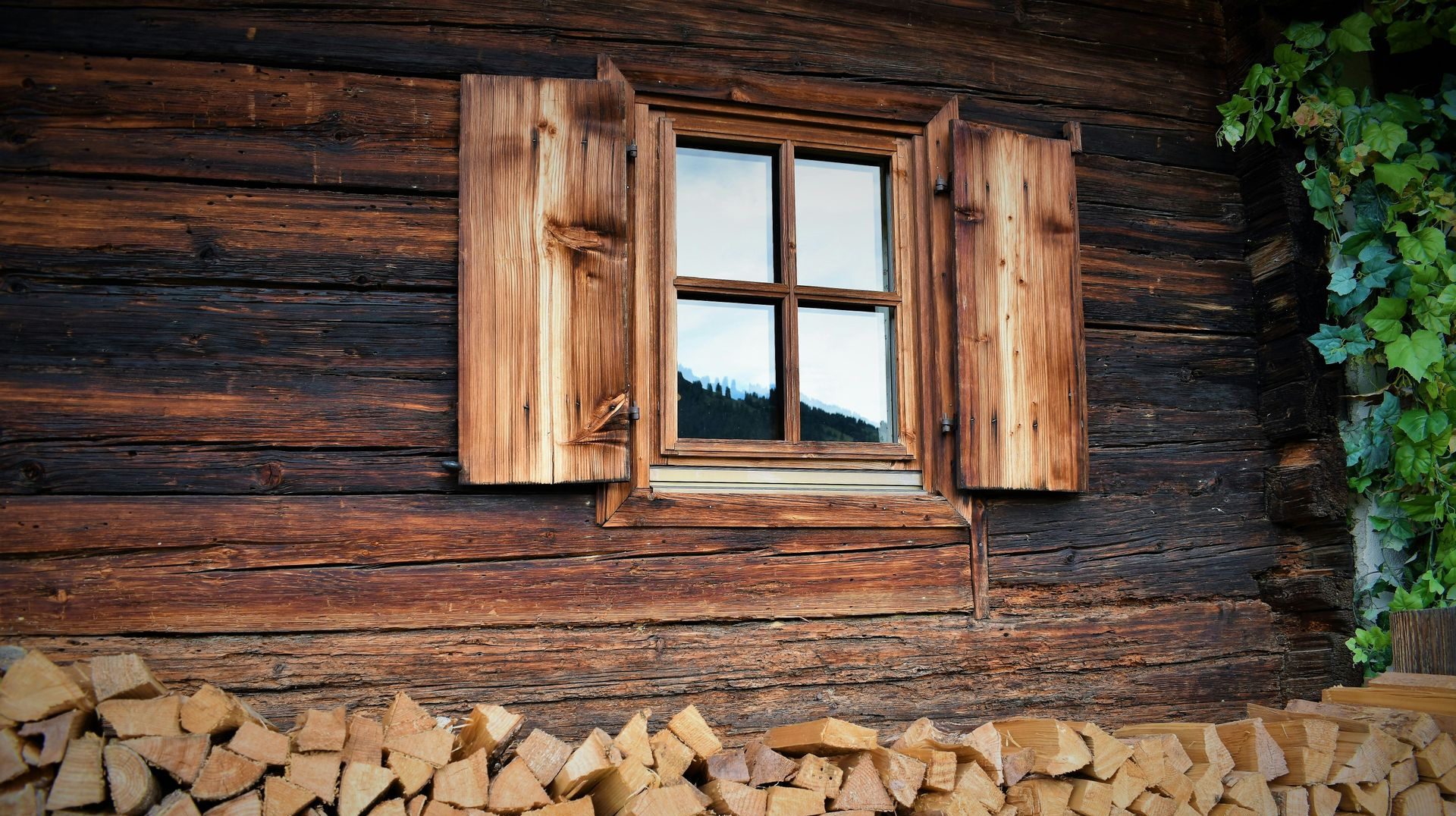 A weathered wooden cabin wall with a small window, open shutters, and a large stack of chopped firewood at the base.