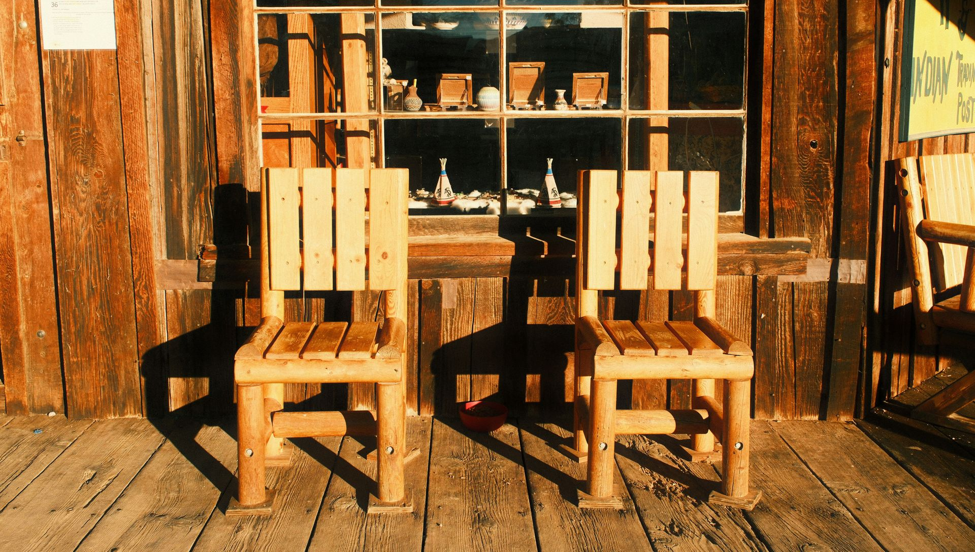 Two wooden chairs on a rustic porch in front of a shop window with items on display, bathed in warm sunset light.