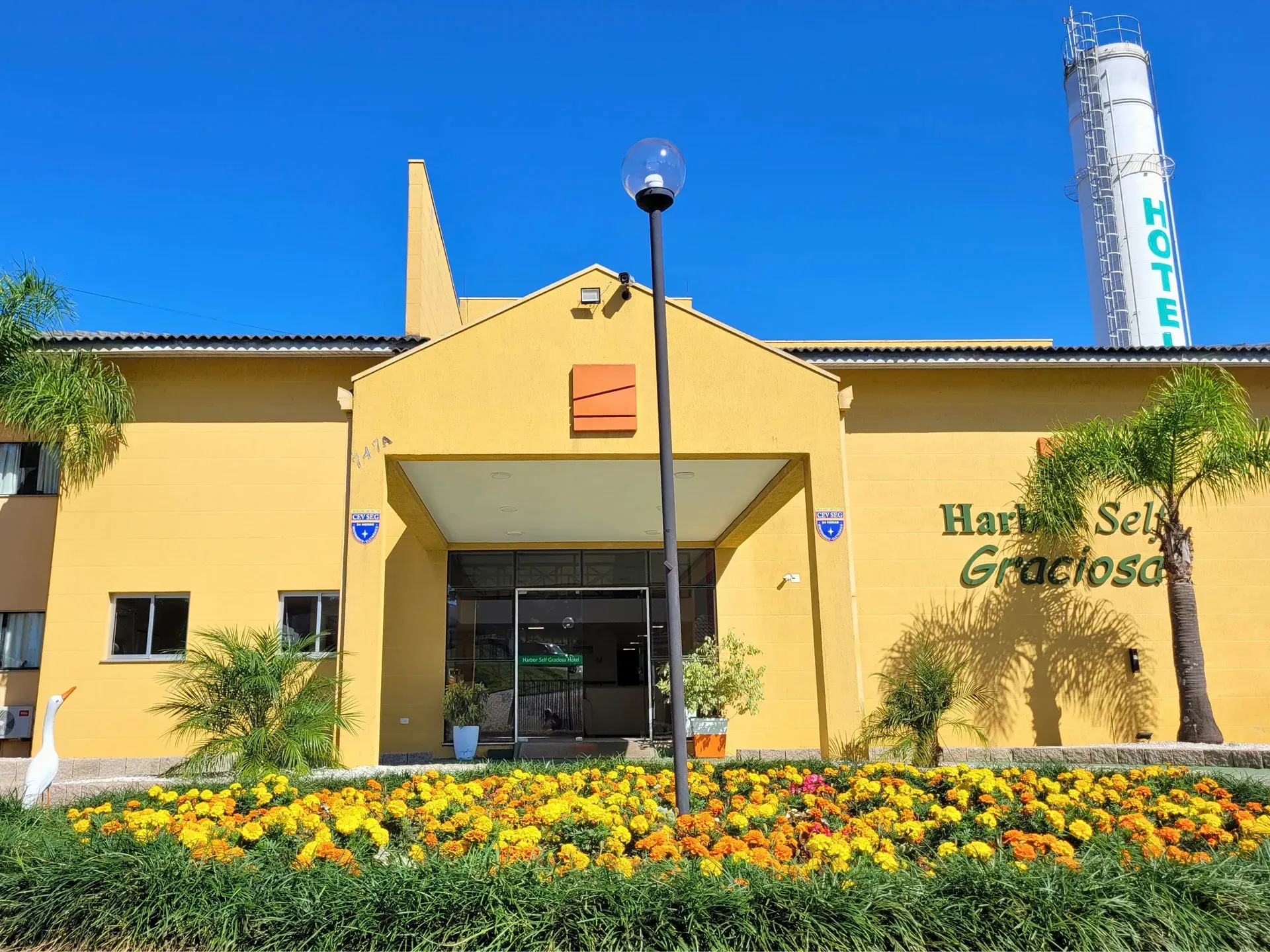 Entrada amarela do hotel com portas de vidro, canteiros de flores e céu azul. Nome do hotel à direita.