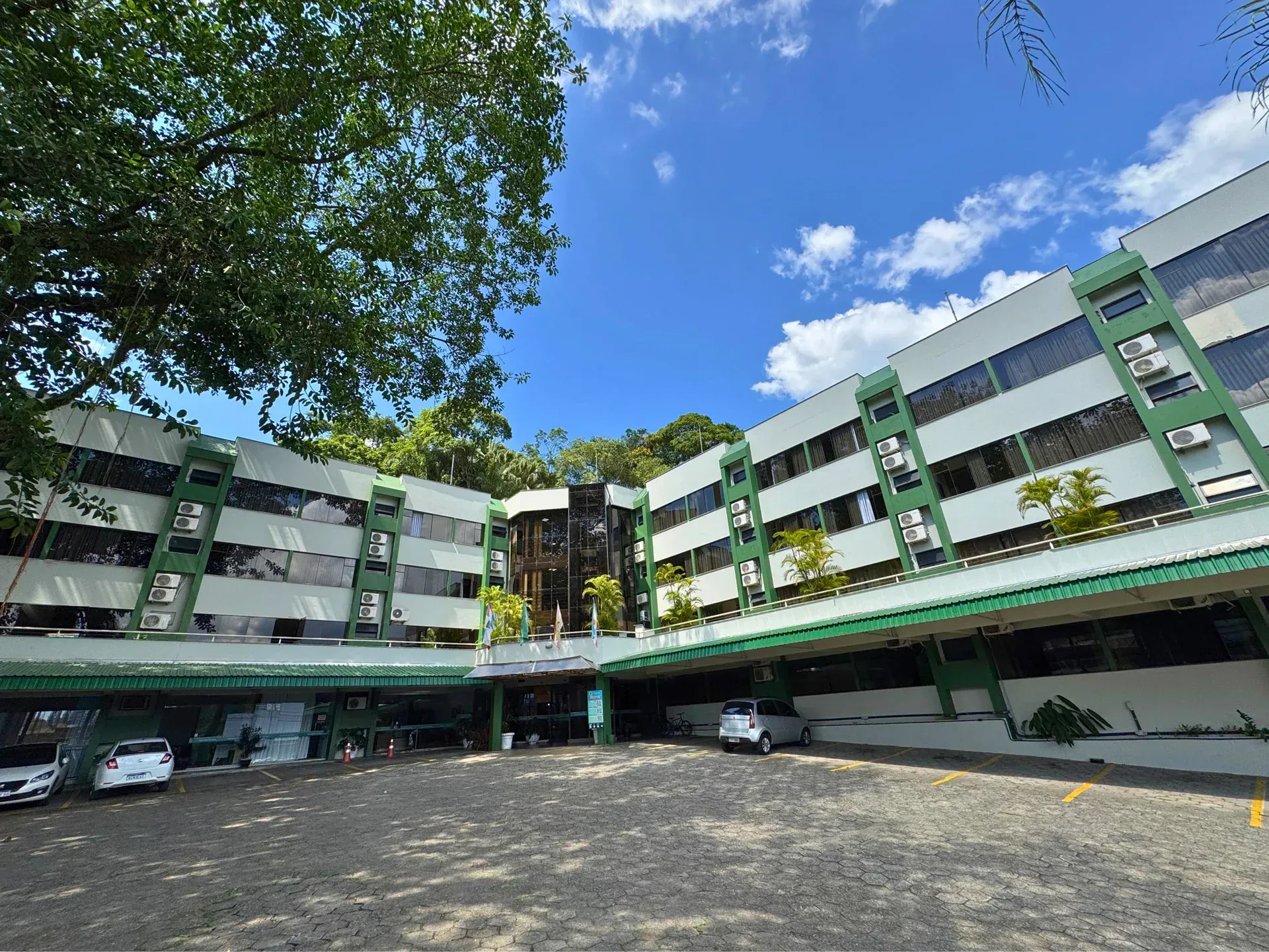 Prédio de vários andares com detalhes em verde, carros estacionados, árvores e céu azul.