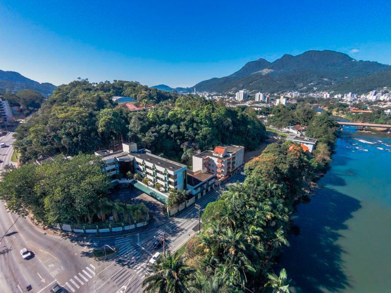 Prédio azul e branco com um amplo estacionamento, palmeiras e uma rua em frente. Dia ensolarado.