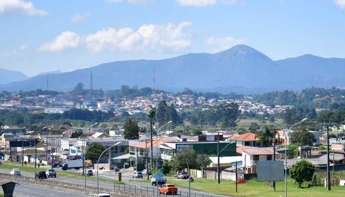 Cidade com prédios, árvores e estradas. Montanhas ao fundo sob um céu azul com nuvens.