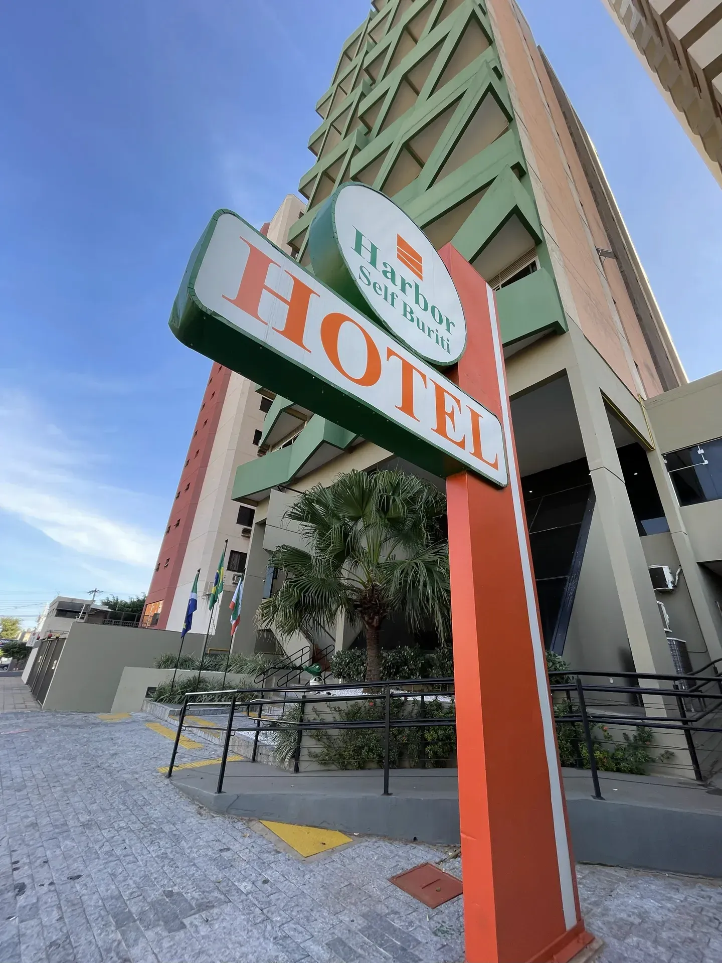 Prédio azul e branco com um amplo estacionamento, palmeiras e uma rua em frente. Dia ensolarado.