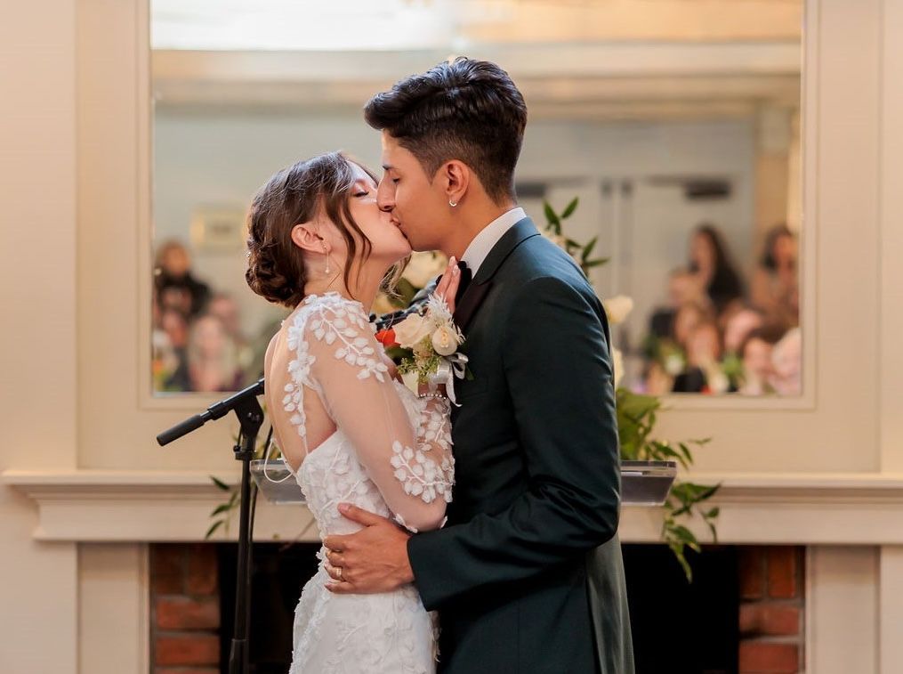 A bride and groom are kissing in front of a fireplace.