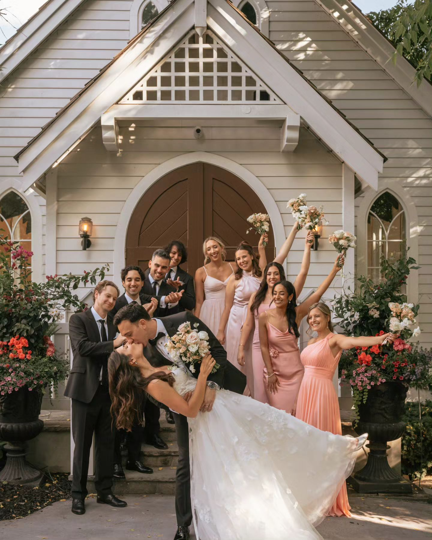 A bride and groom are kissing in front of a church with their wedding party.