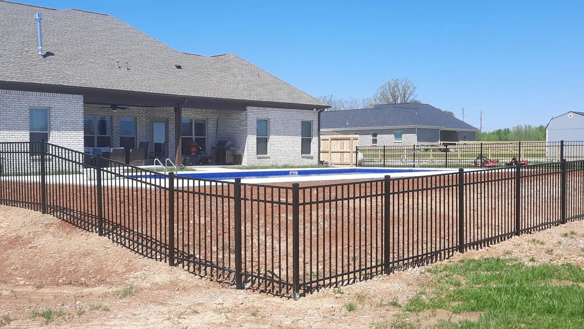 A black metal fence encloses a backyard swimming pool behind a suburban brick home under a clear blue sky.