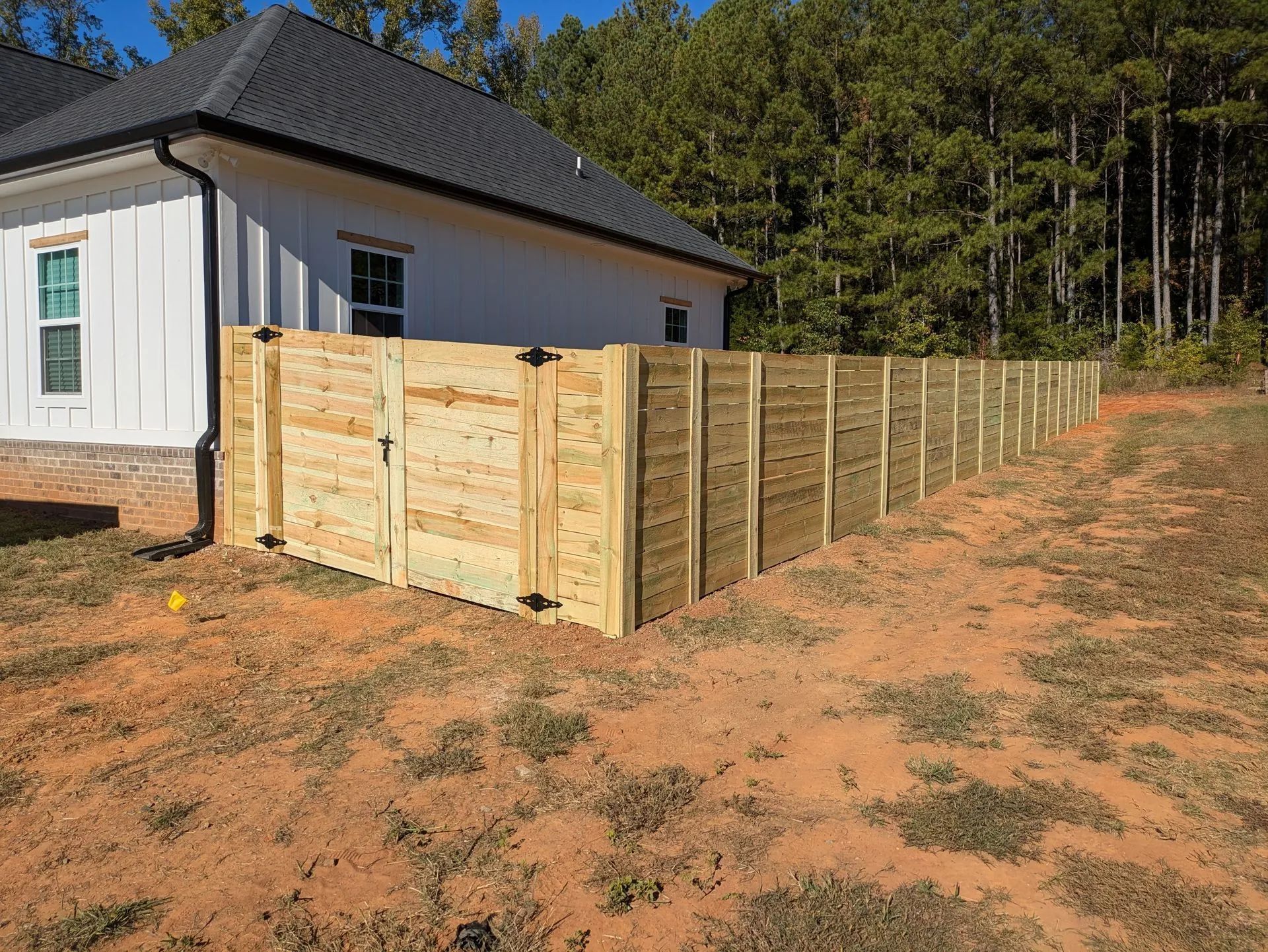 Wooden fence around a house, on a brown yard with a background of trees.