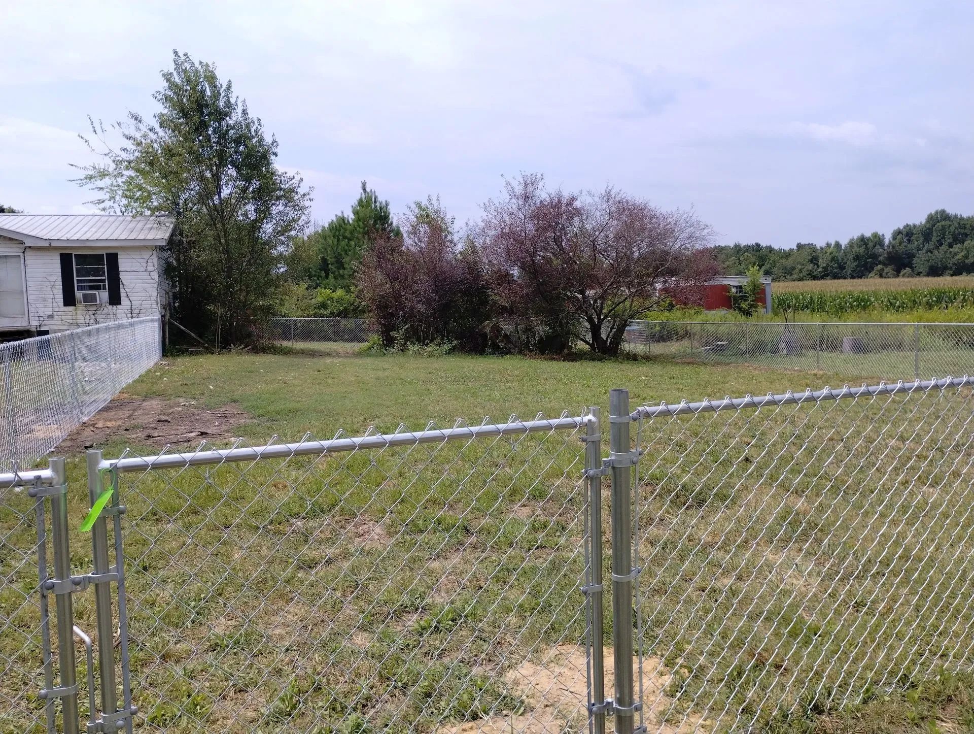 Chain-link fence in a grassy yard, partially framing a mobile home and shrubs, with fields and trees in the distance.