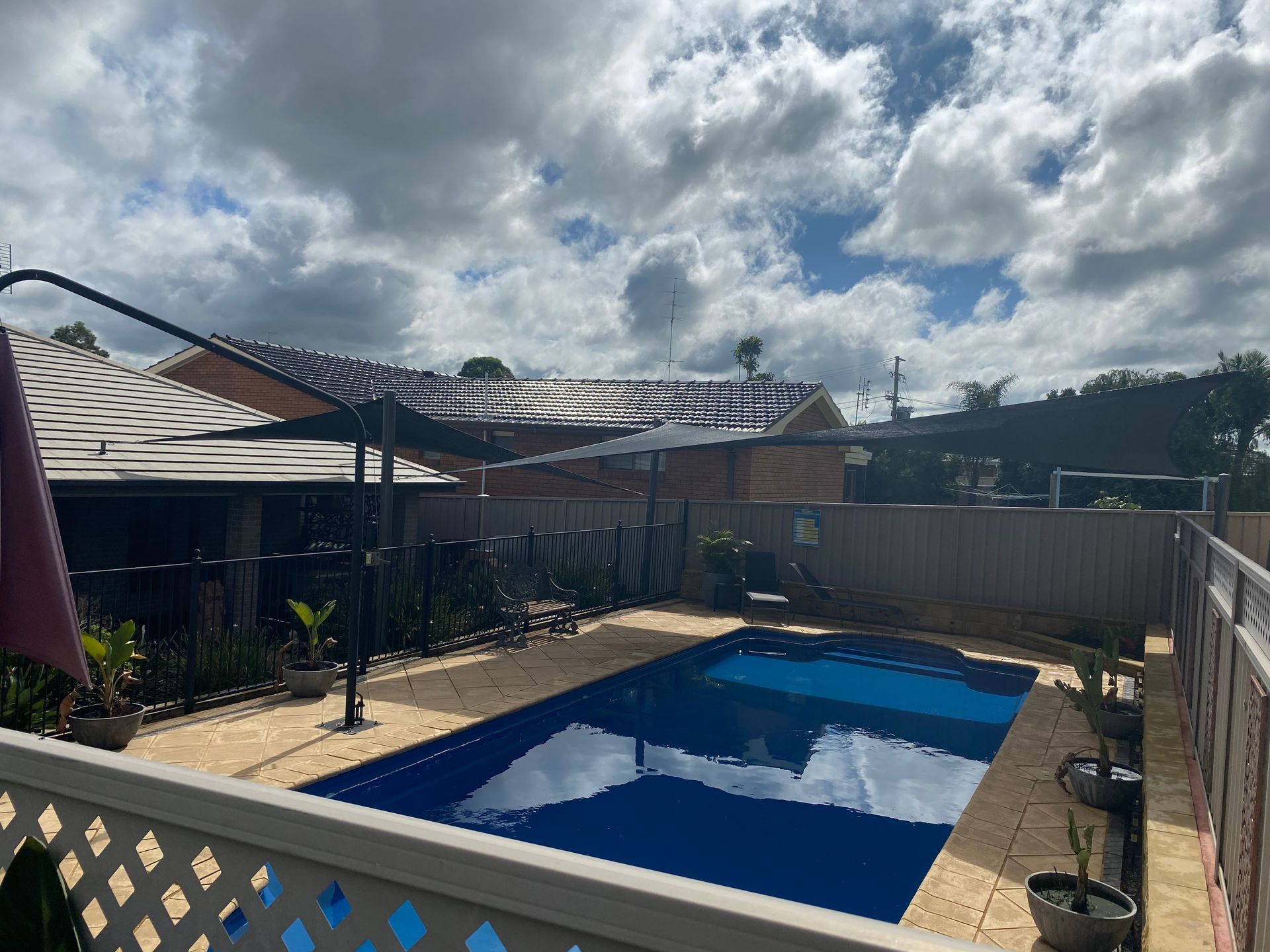 A rectangular swimming pool with deep blue water set in a stone patio with a shade sail, backyard fence, and house — Shades of Blue In North Wyong, NSW