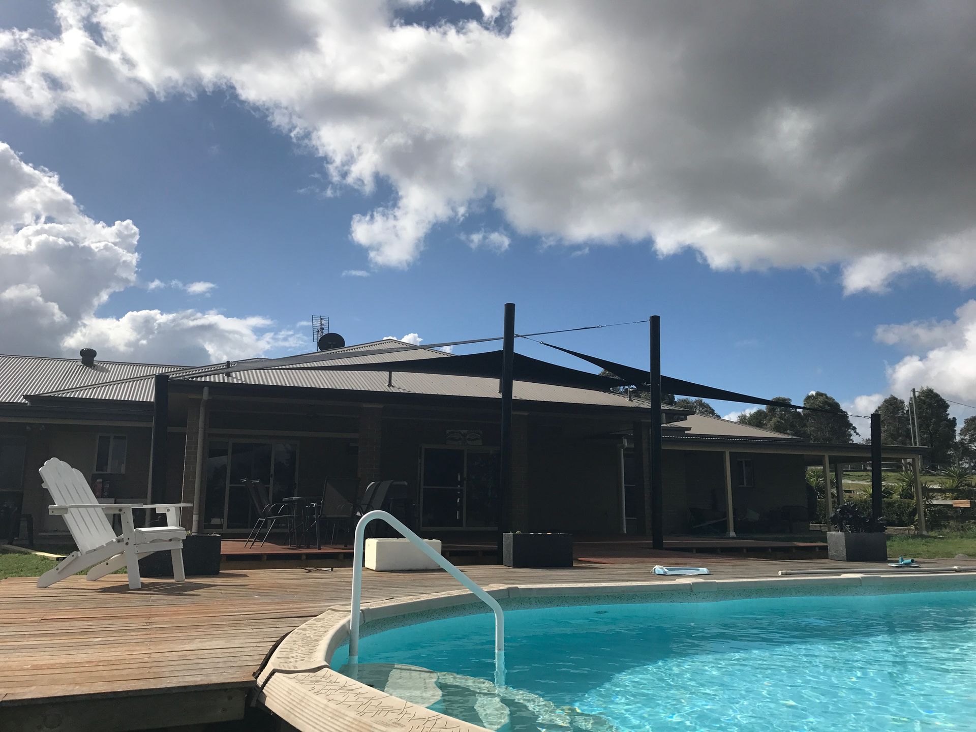 A swimming pool with a white chair on the deck, facing a patio area with shade sails under a blue, cloudy sky — Shades of Blue In North Wyong, NSW