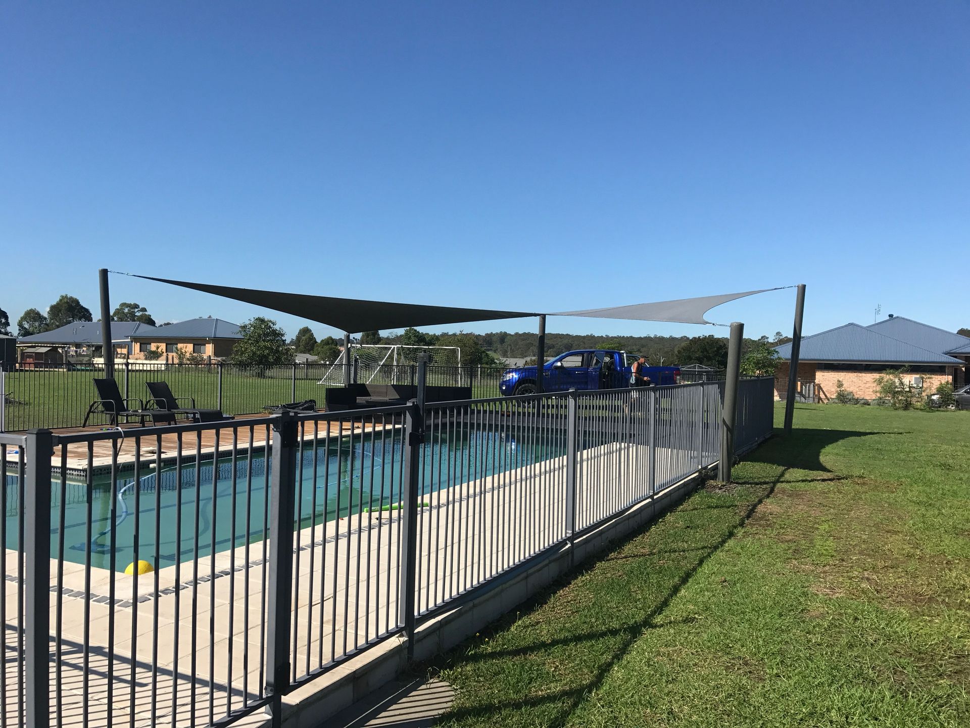 An outdoor swimming pool enclosed by a dark metal fence, covered by a large triangular shade sail on a sunny day — Shades of Blue In North Wyong, NSW