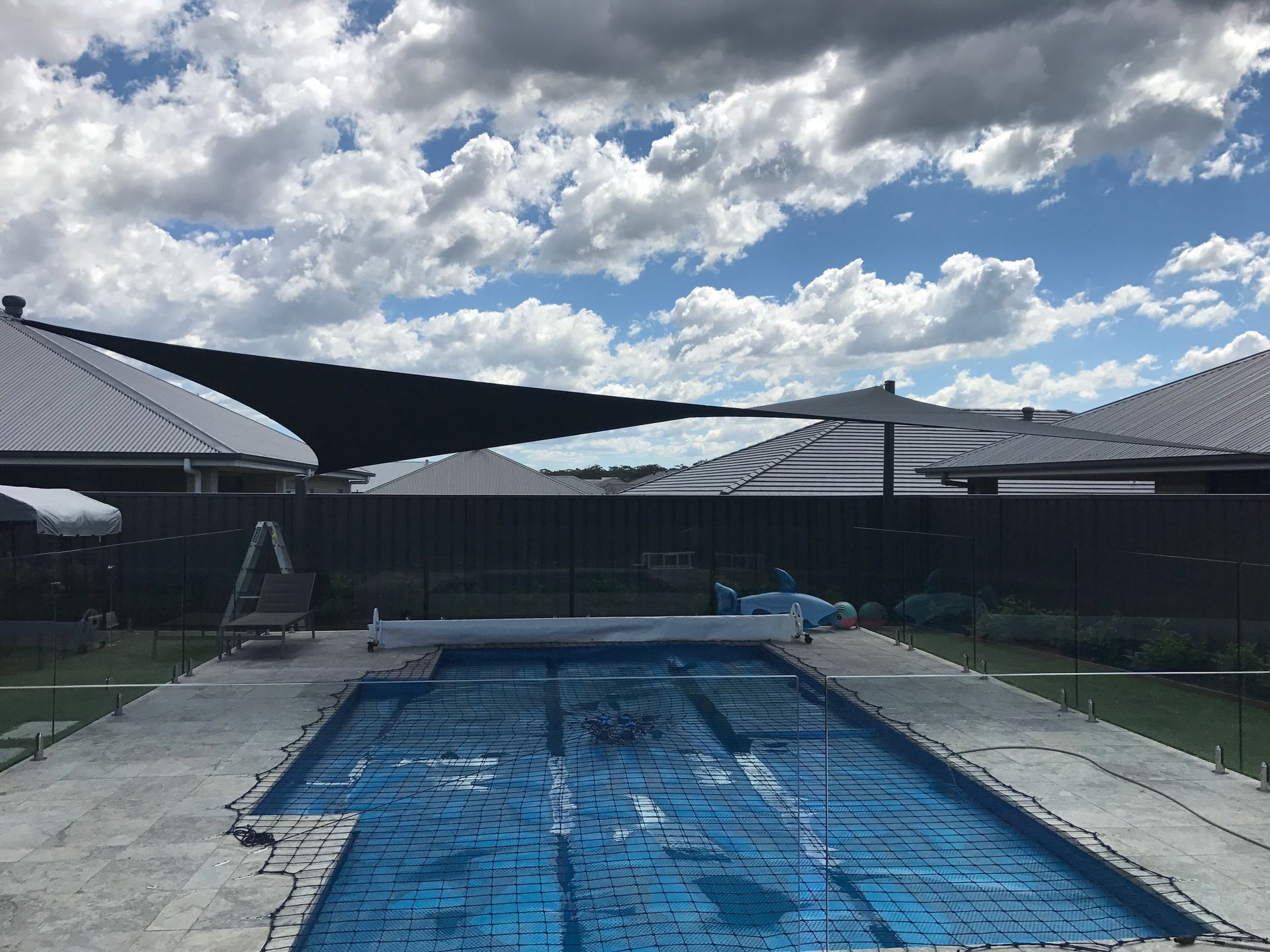 A rectangular swimming pool covered with a blue tarp in a backyard with a large black shade sail and a fence  — Shades of Blue In North Wyong, NSW