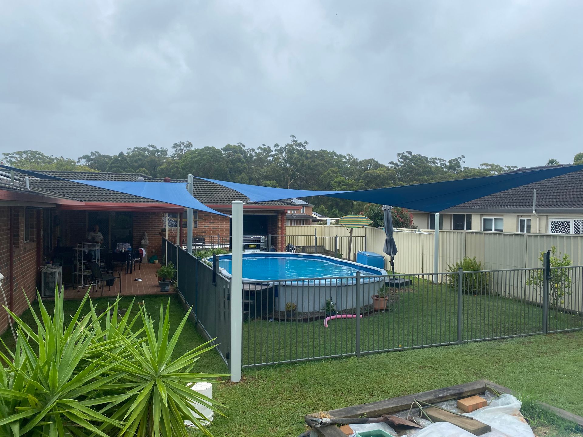 A backyard swimming pool shaded by a large, dark triangular sail canopy under a sunny blue sky — Shades of Blue In North Wyong, NSW