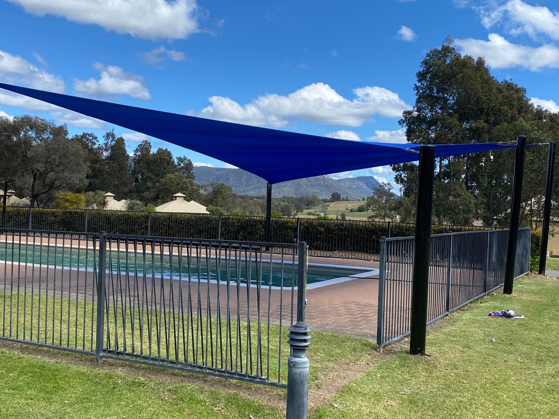 A large blue triangular shade sail mounted on black poles over a backyard swimming pool in a sunny, grassy area— Shades of Blue In North Wyong, NSW