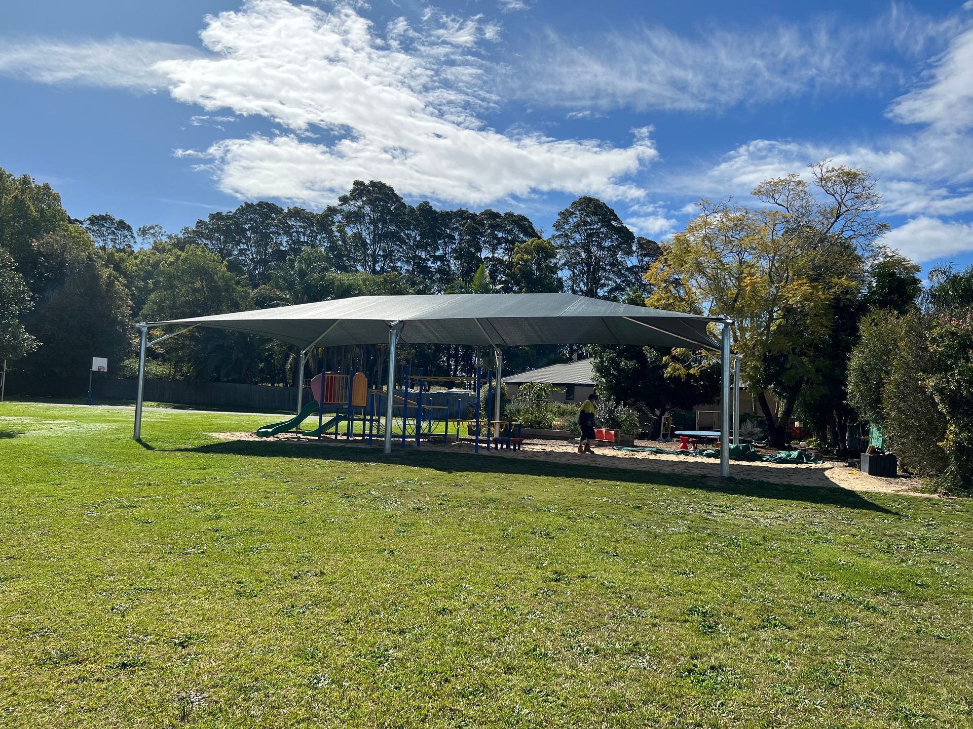 A playground under a large shade structure on a grassy lawn with trees in the background under a blue, cloudy sky — Shades of Blue In North Wyong, NSW