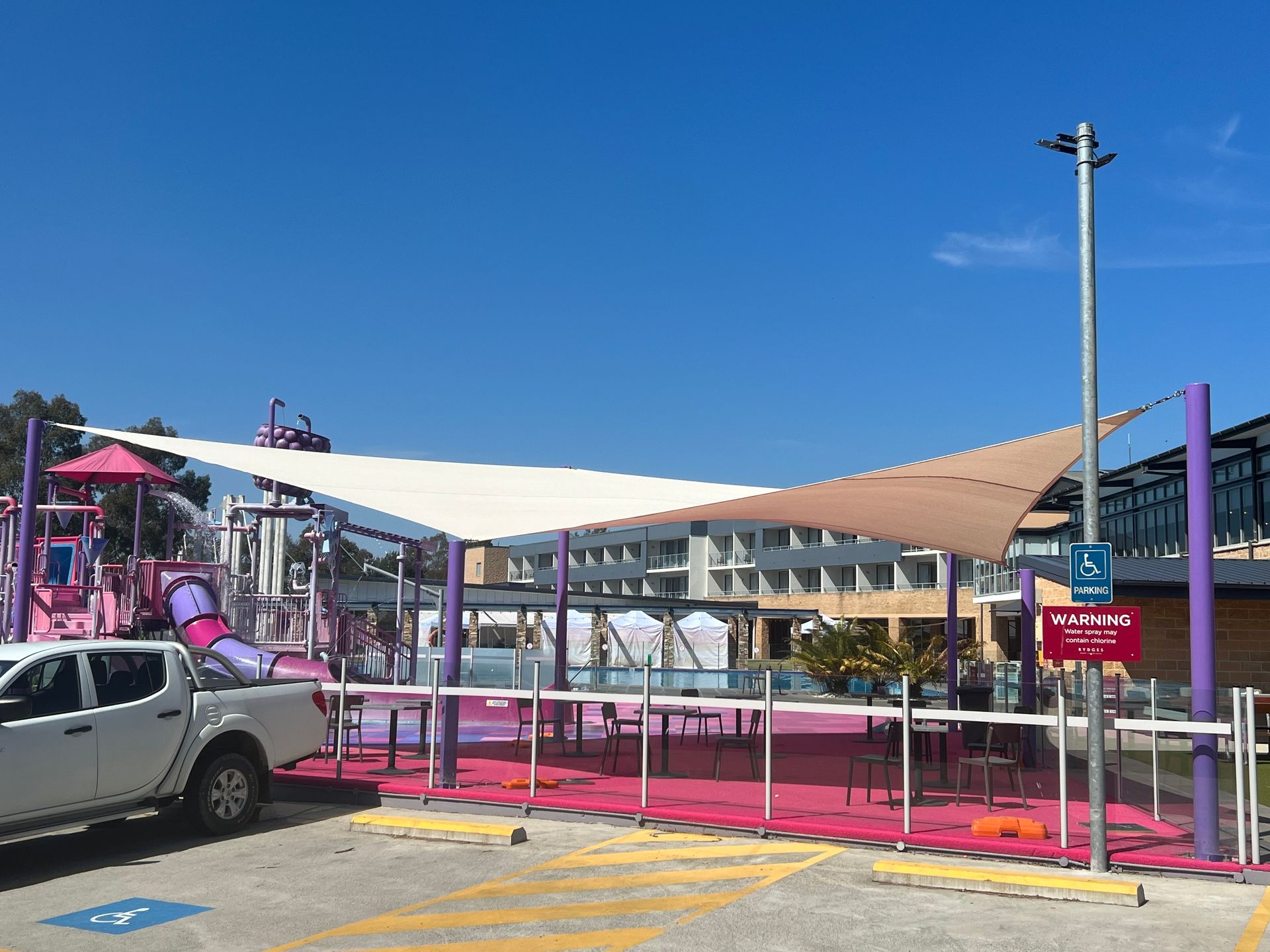 A white, triangular shade sail over a pink-paved area with playground equipment and a parked truck in a sunny lot — Shades of Blue In North Wyong, NSW