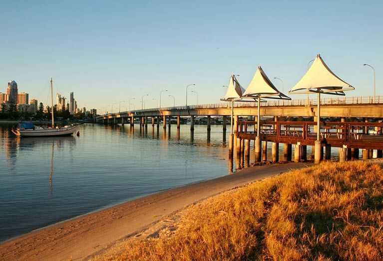 A sunlit sandy shore leads to a wooden pier with three conical white shades, near a bridge and distant city skyline — Shades of Blue In North Wyong, NSW