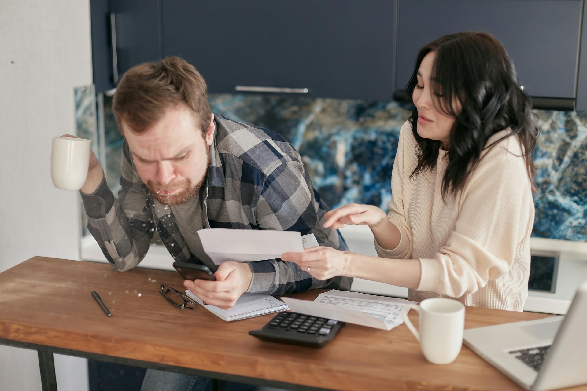Couple reviewing bills in kitchen, man on phone, woman pointing, looking concerned.