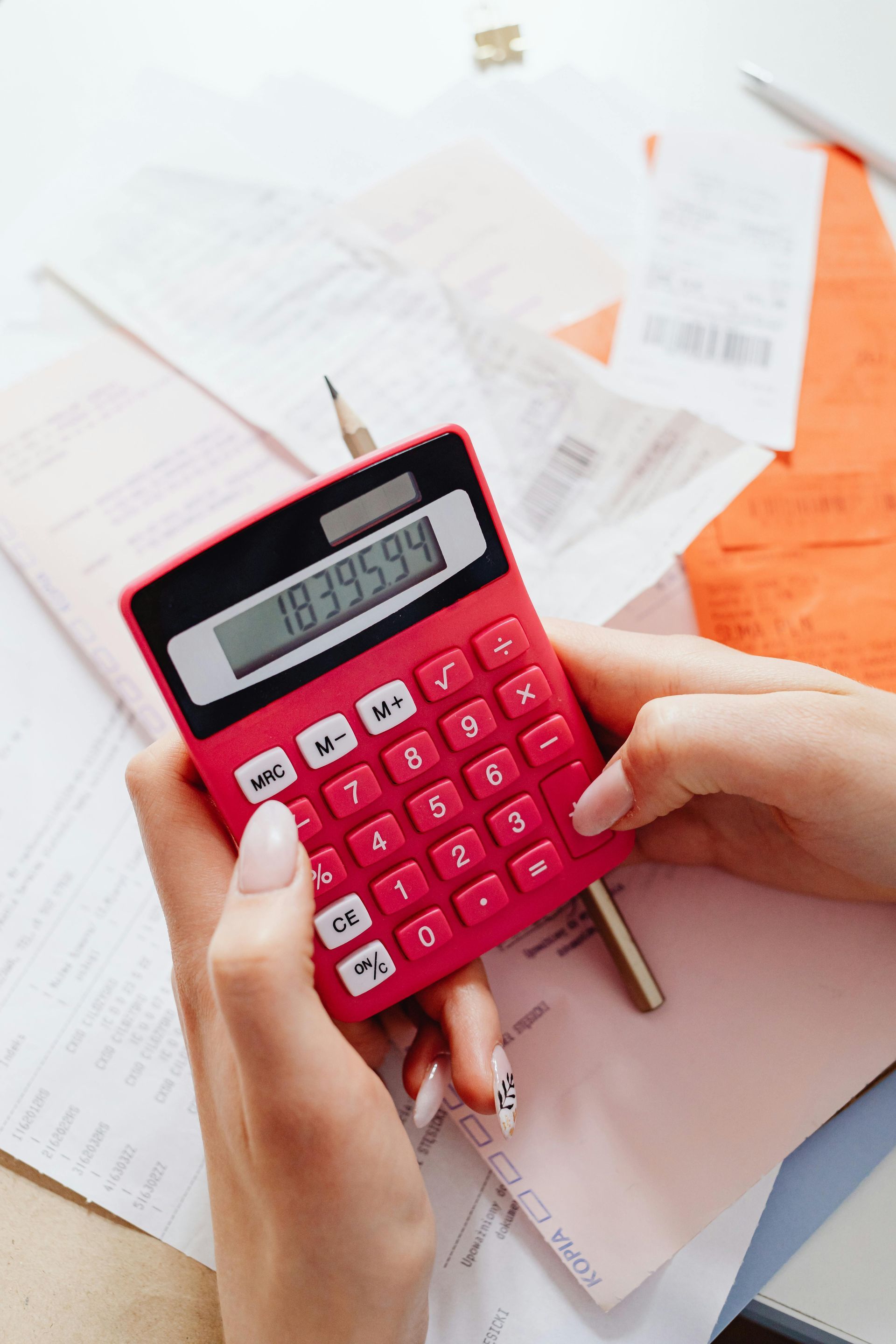 Hands using a red calculator with a display of 18395544. Receipts and papers are scattered around.