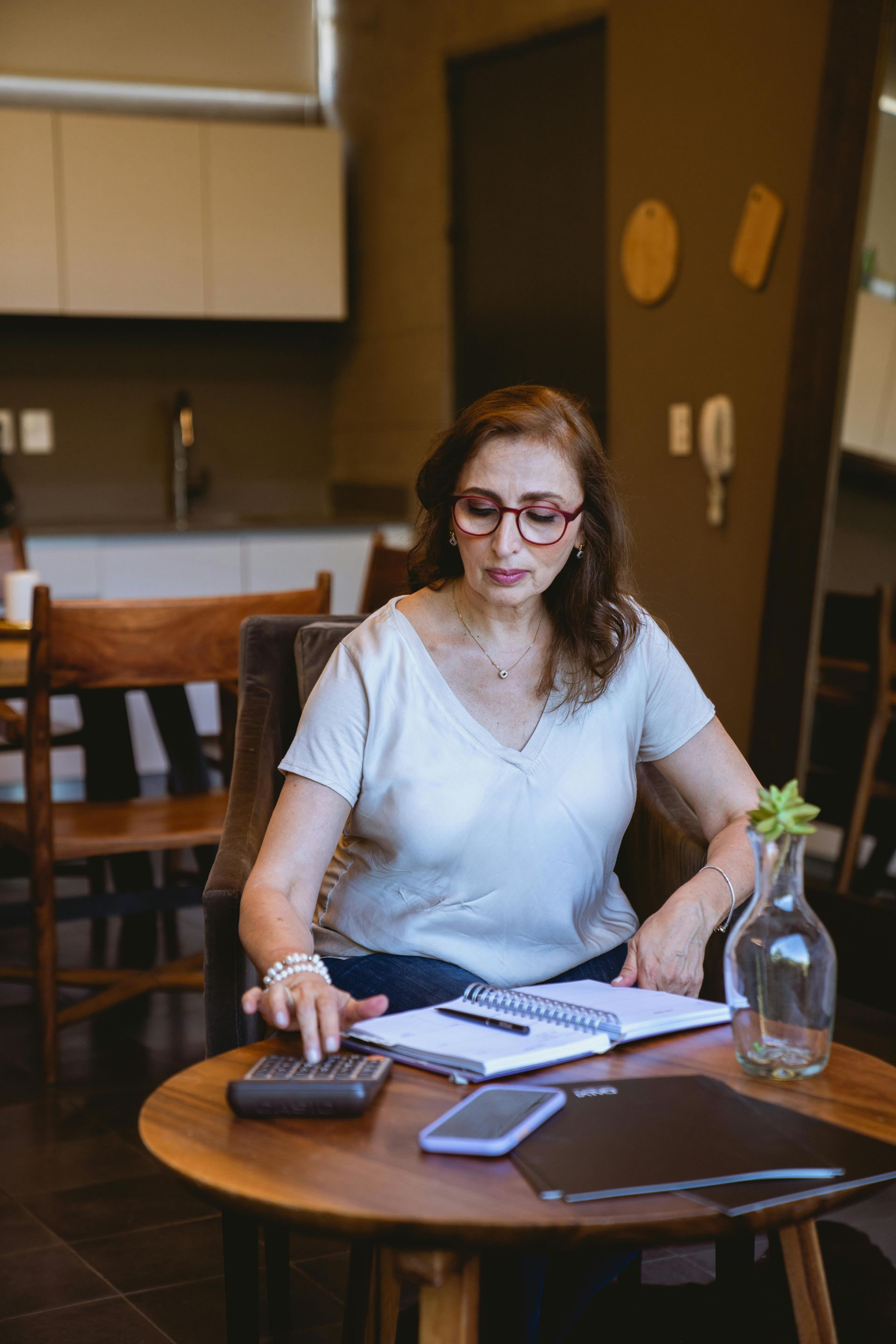 Woman with glasses using a calculator at a table with notebook, phone, and laptop in a cafe.