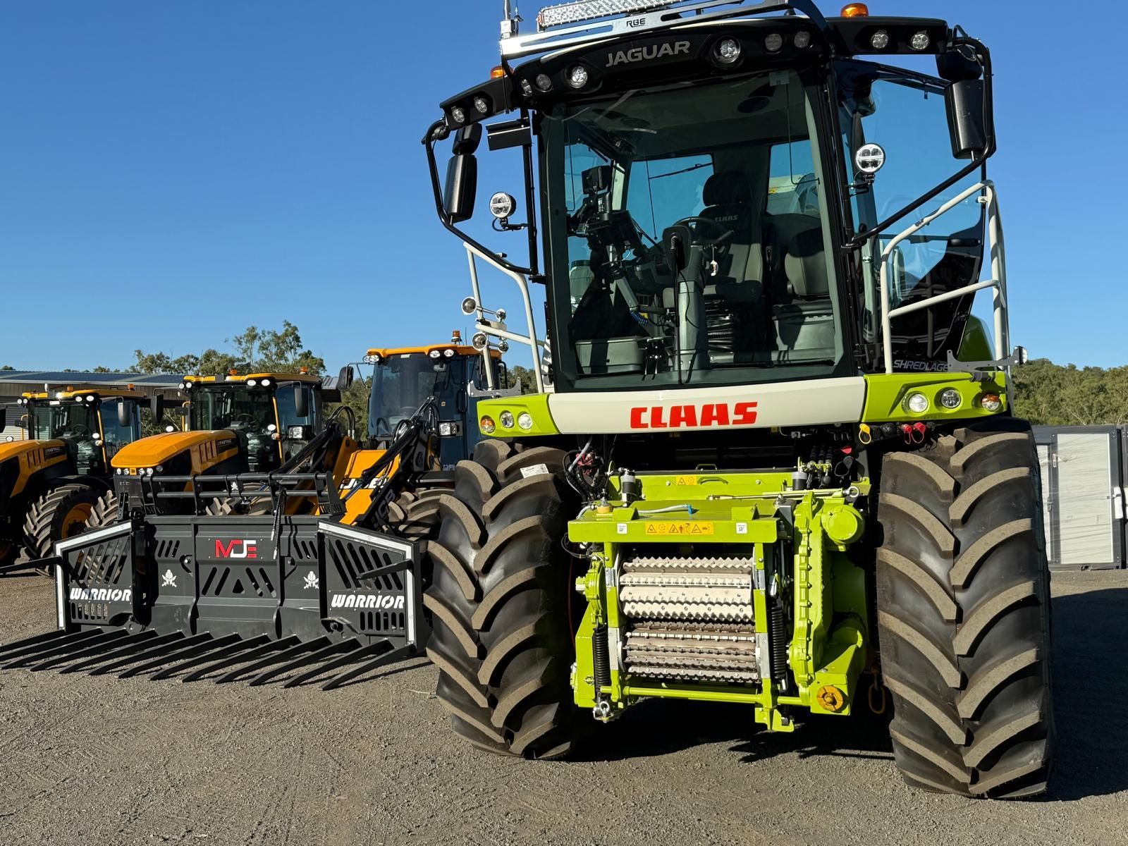 A lime green and white CLAAS forage harvester parked on gravel with other farm equipment in the background.