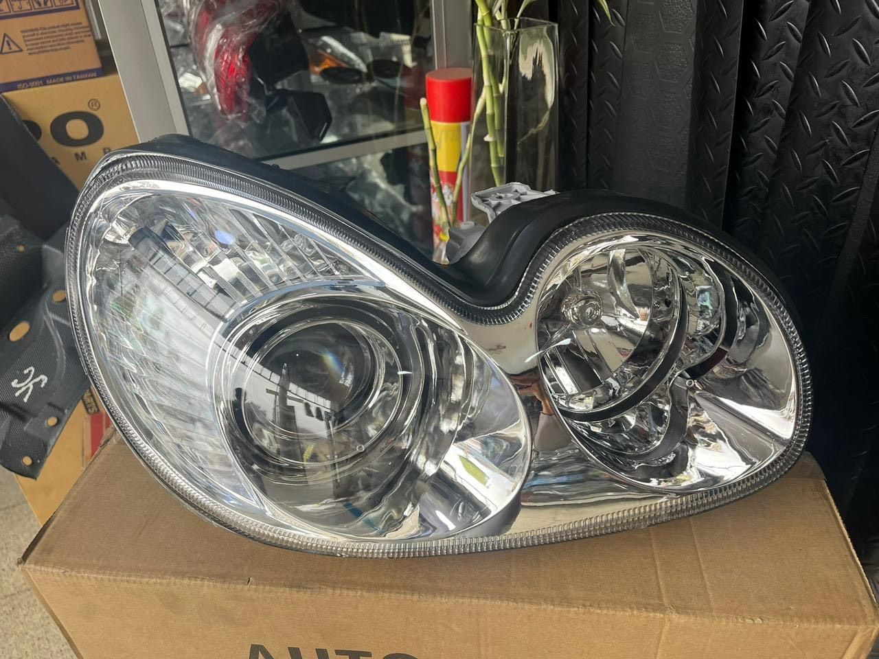 Faro de coche, cromado y transparente, sobre caja de cartón.