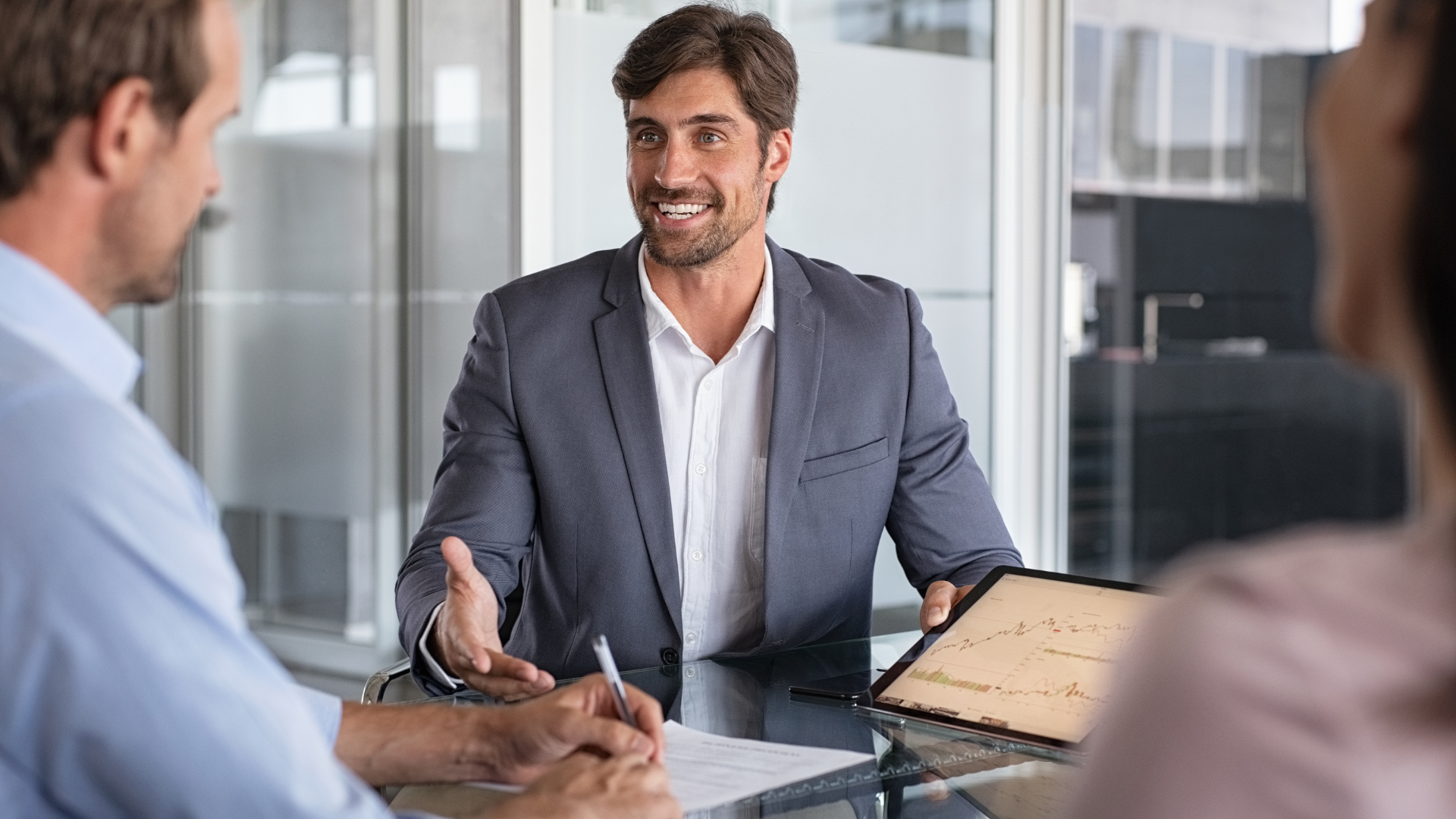 A man in a suit is sitting at a table talking to two people.