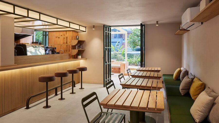 Cafetería interior con barra iluminada, taburetes modernos y mesas de madera, conectada al exterior.