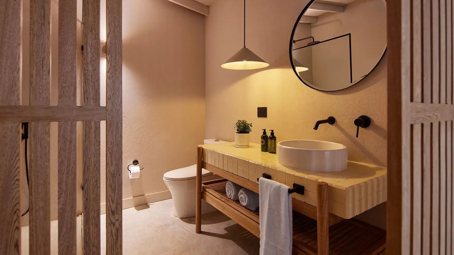 Lavamanos moderno en el baño de la habitación de lujo Rinja, con espejo redondo, decoración en madera y elegante iluminación, proporcionando una experiencia de confort y sofisticación.