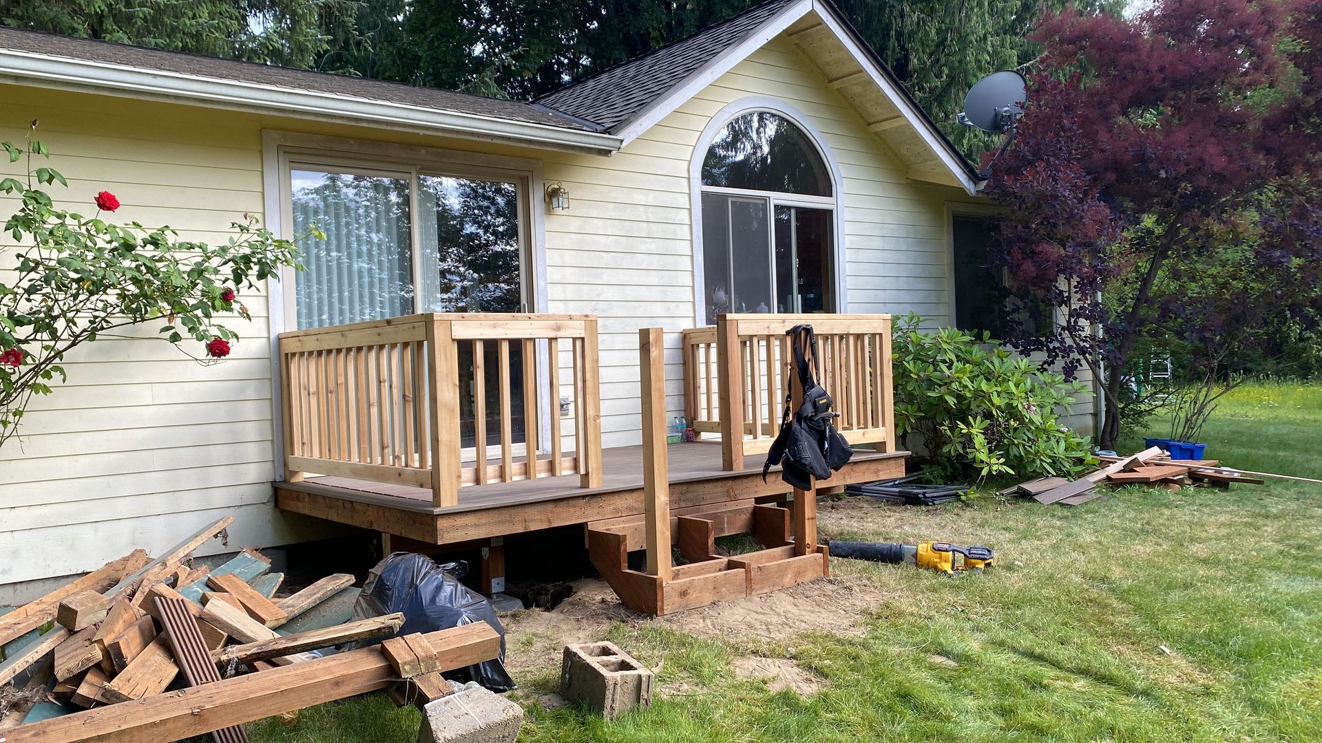 Wooden deck under construction next to a yellow house. Bricks and tools scattered in yard.