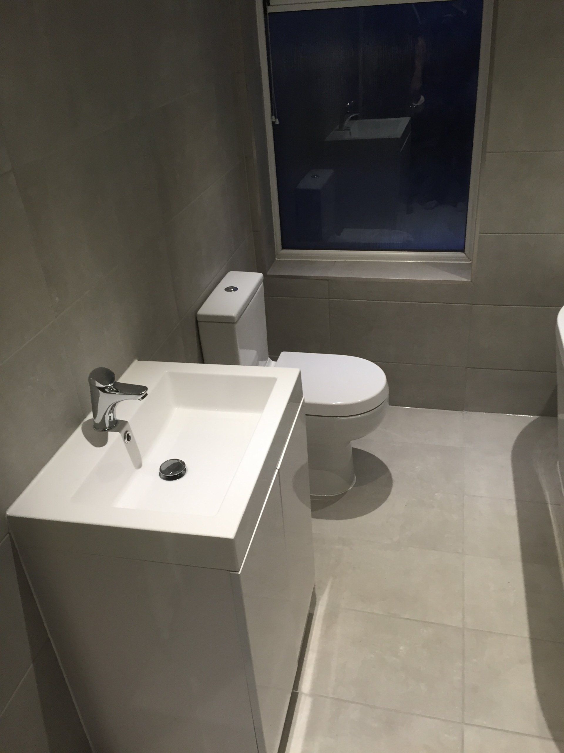 Modern bathroom with white sink, toilet, and gray tiled walls. A window is in the background.