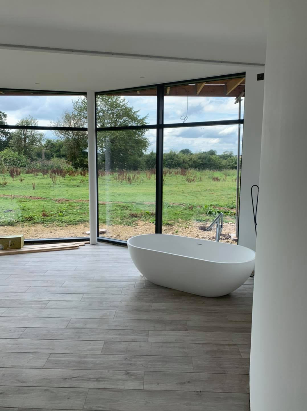 White bathtub in a room with large windows overlooking a field. Wooden floor.