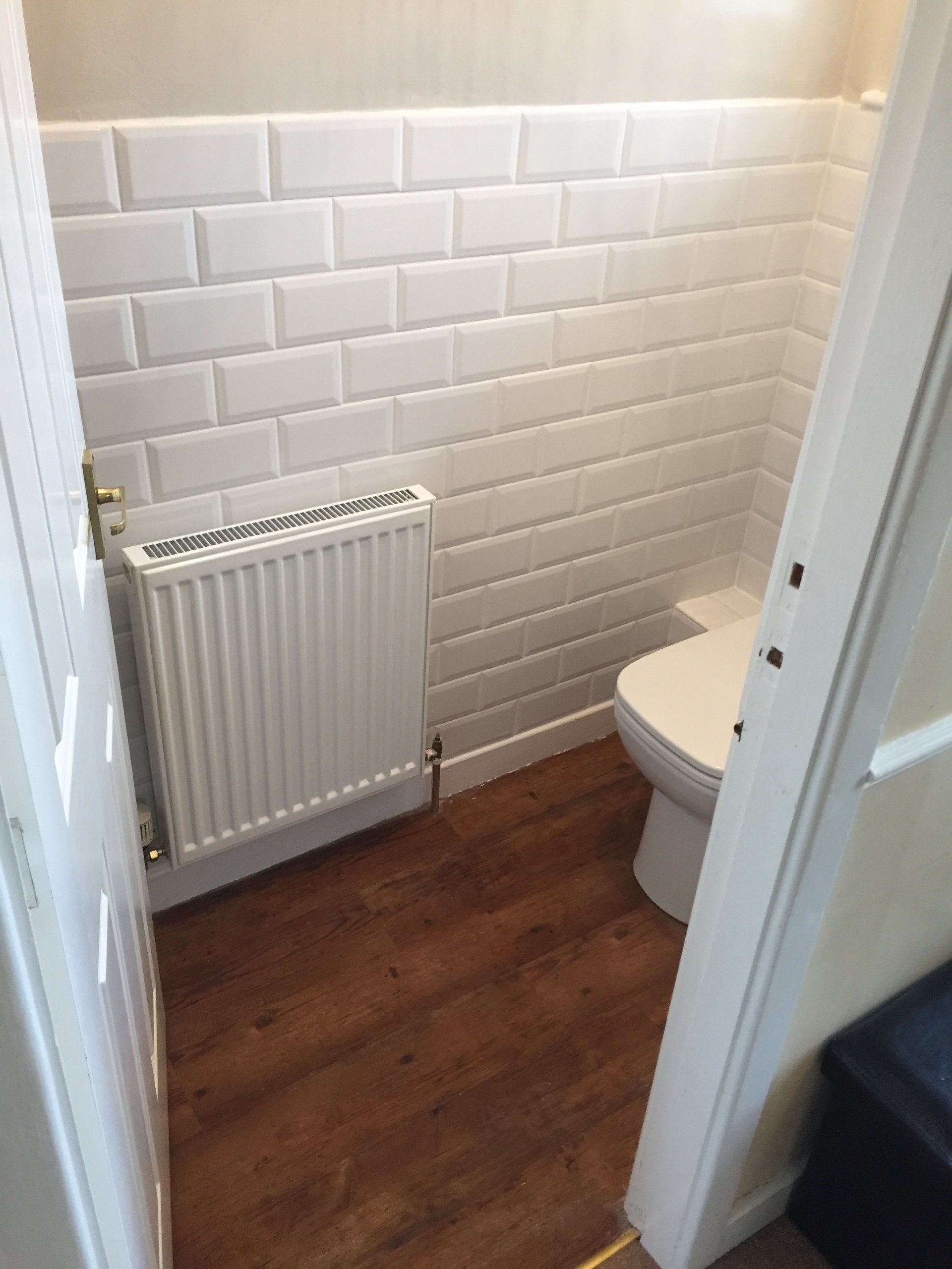 Small white-tiled bathroom with toilet and radiator; wood-look flooring. Partially open white door.