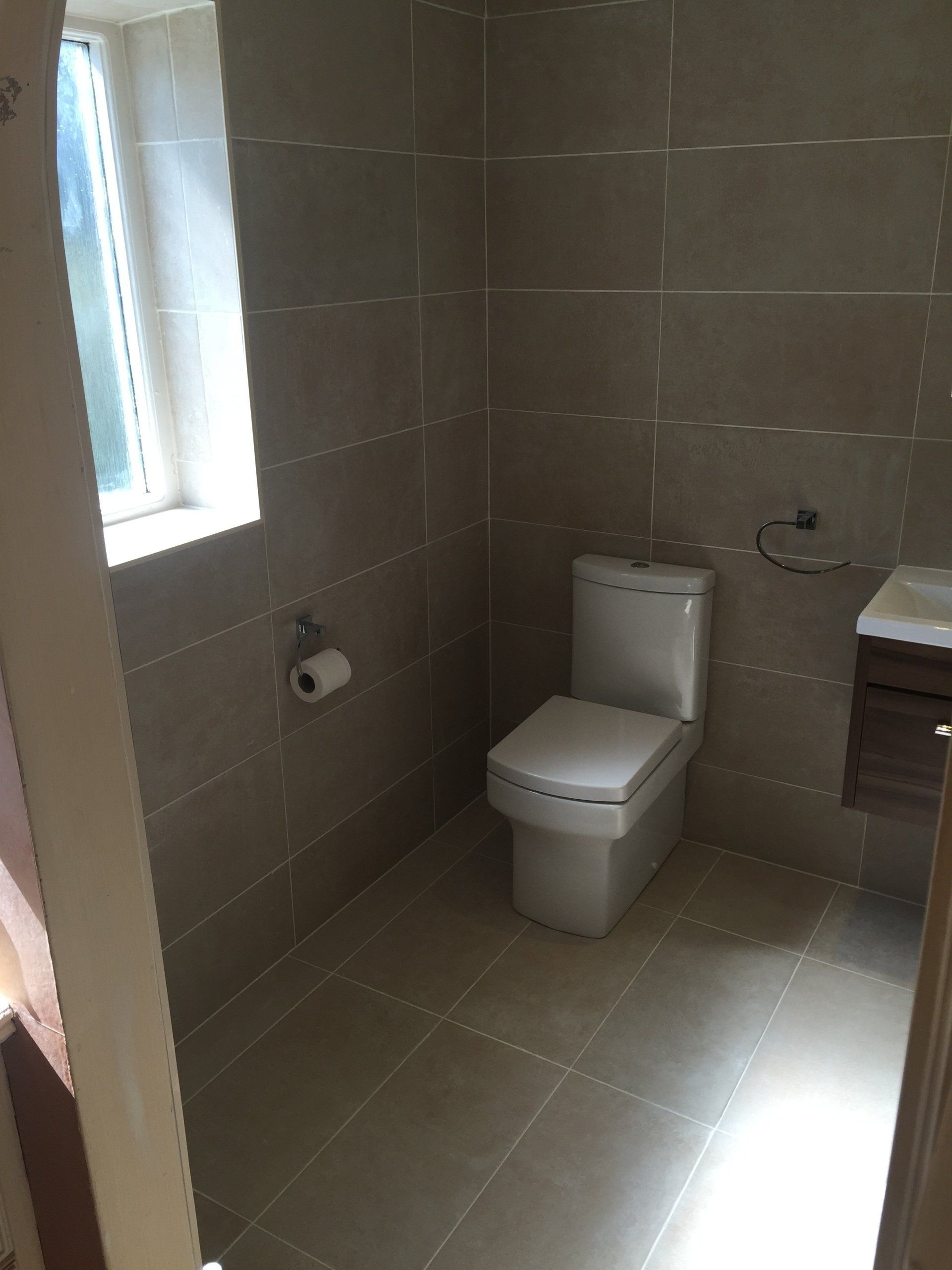 A small bathroom with tiled walls and floor. It features a toilet, a small window, and a vanity.