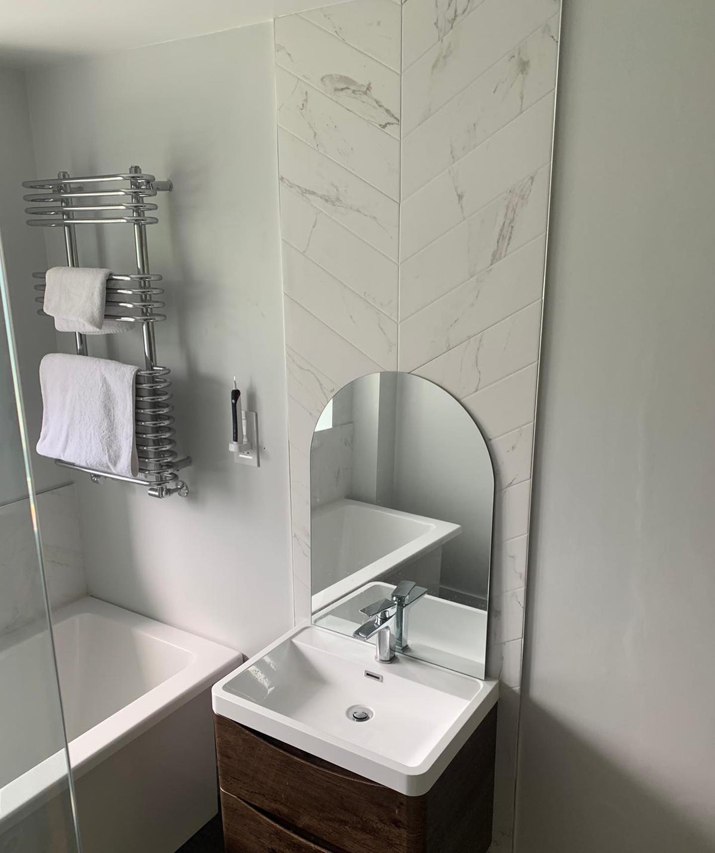 Modern bathroom with a white tub, sink, and decorative wall tiles. Chrome towel rack and arched mirror.