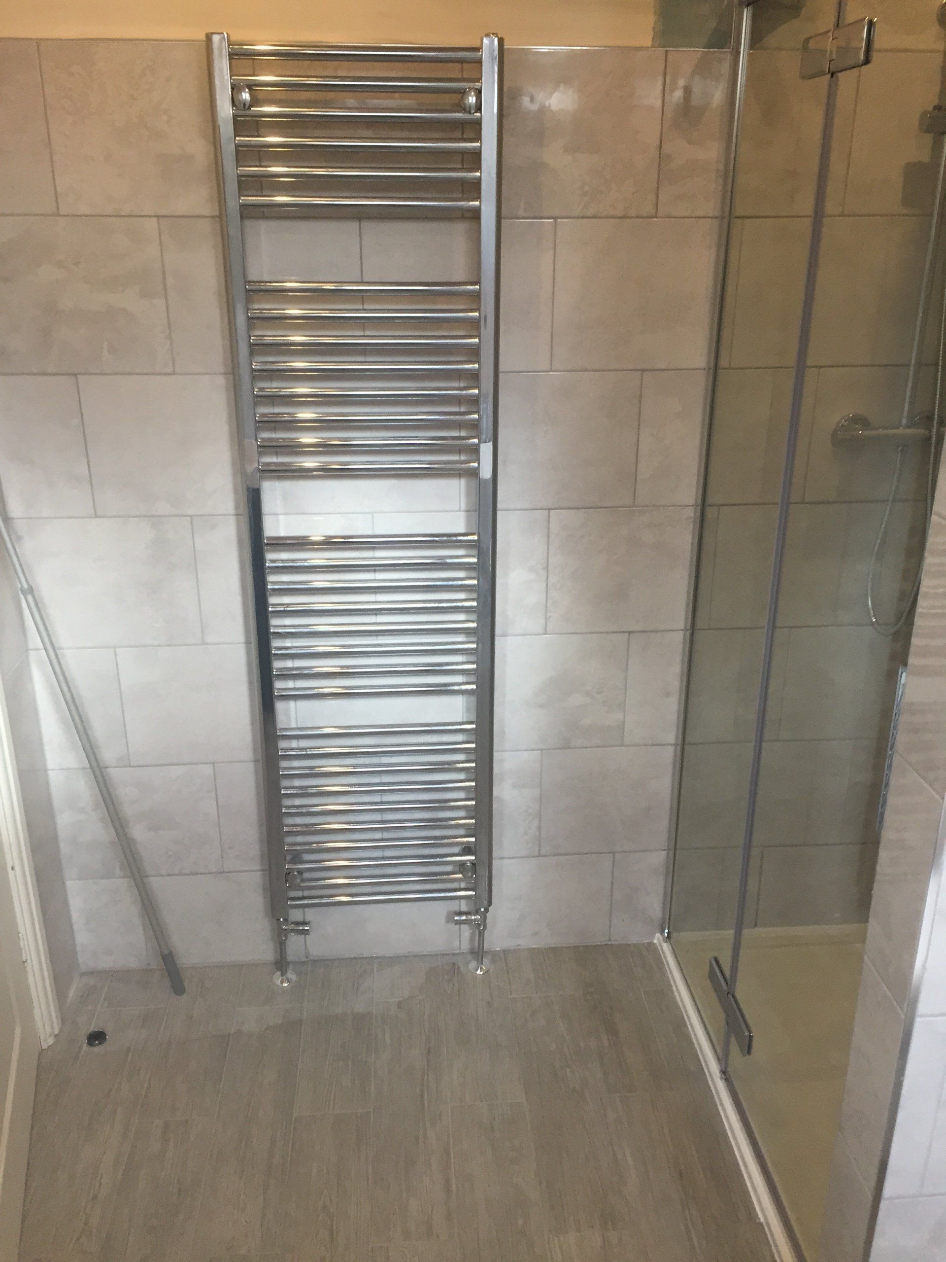 Chrome towel warmer on tiled wall, next to a glass shower screen in a bathroom.