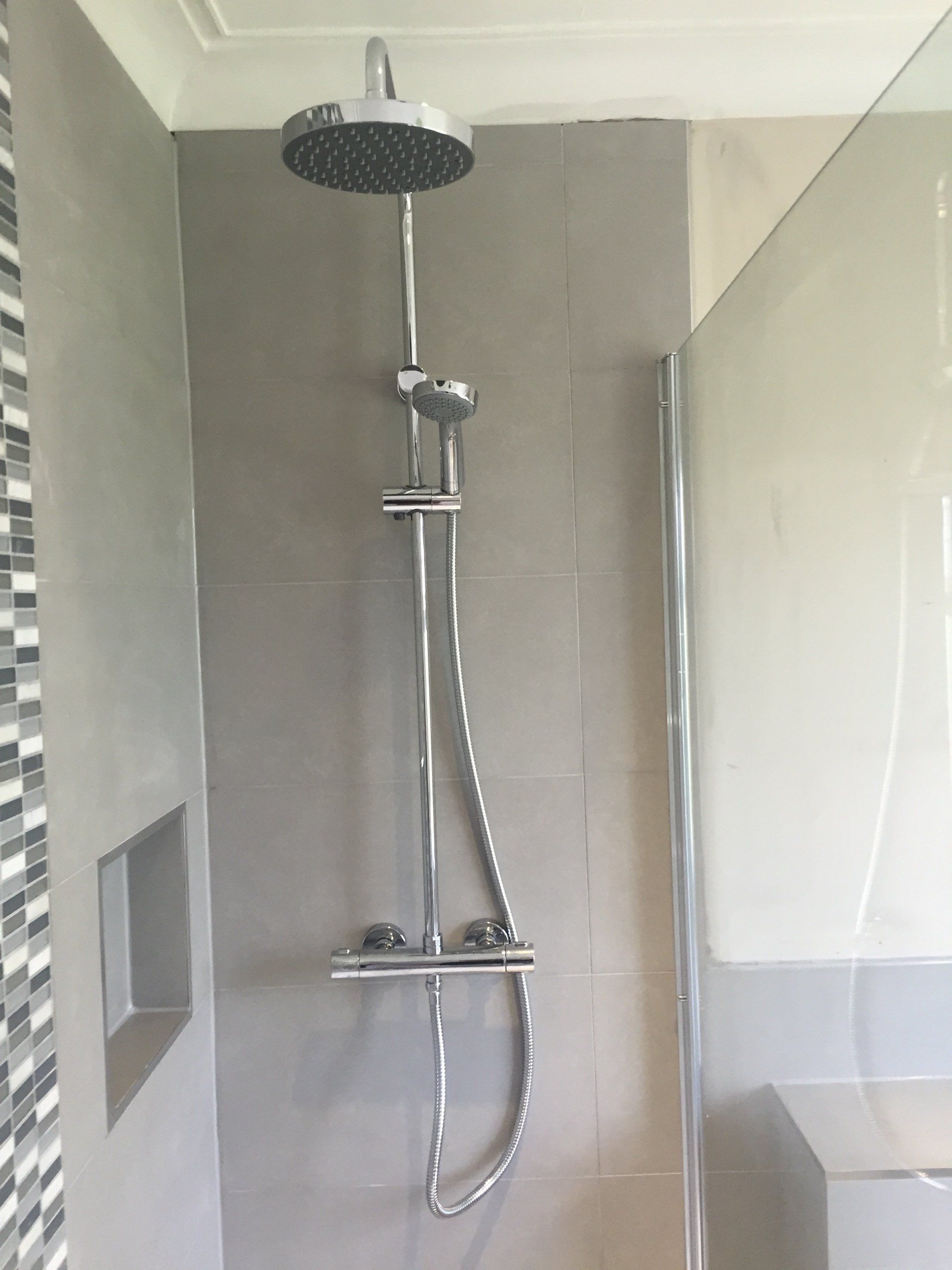 Shower with a large rain head and handheld showerhead, on gray tiled wall, behind a glass door.