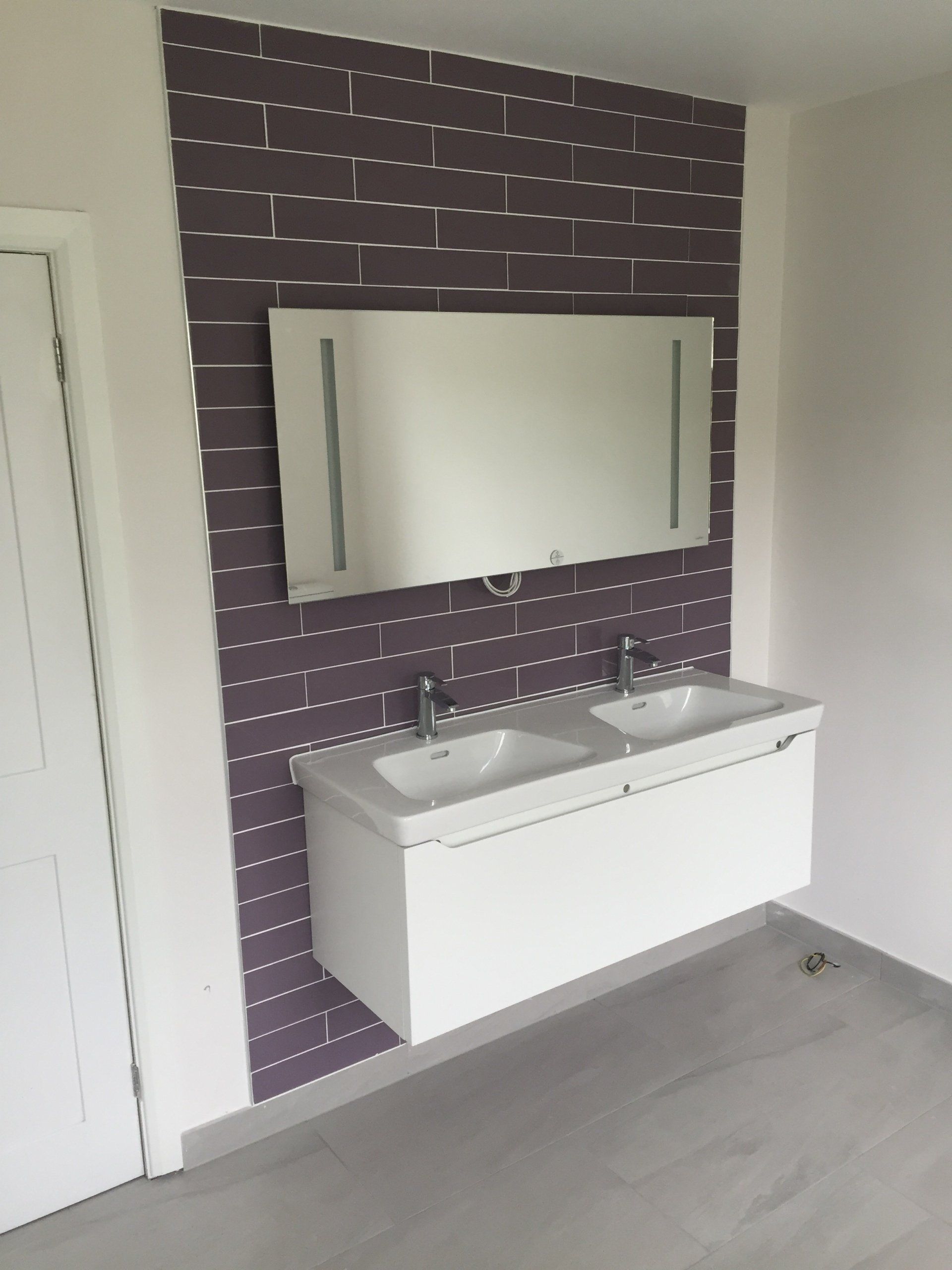 A modern bathroom with a white double sink and mirror, set against a purple brick tiled wall.