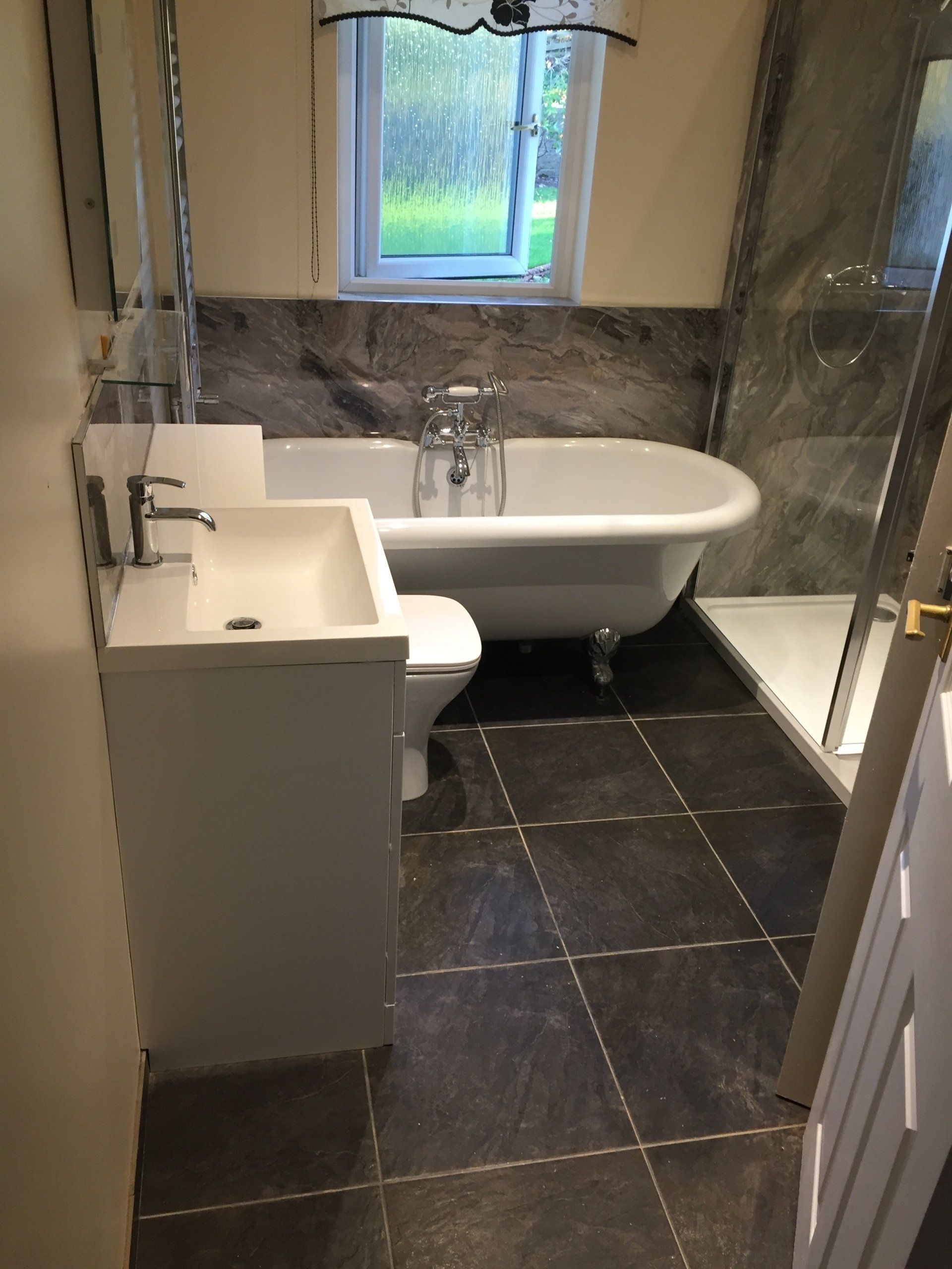 Bathroom with a claw-foot tub, sink, toilet, and shower. Dark tile flooring, marble-look wall tile. Window.