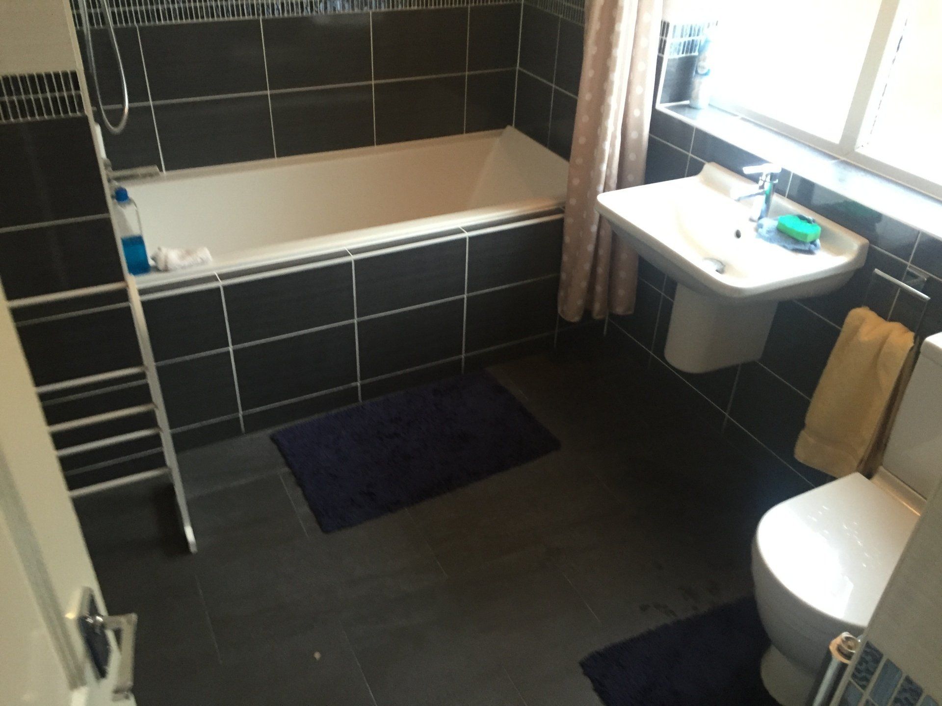 Bathroom with dark gray tiles, white bathtub and sink, toilet, and two dark blue rugs.