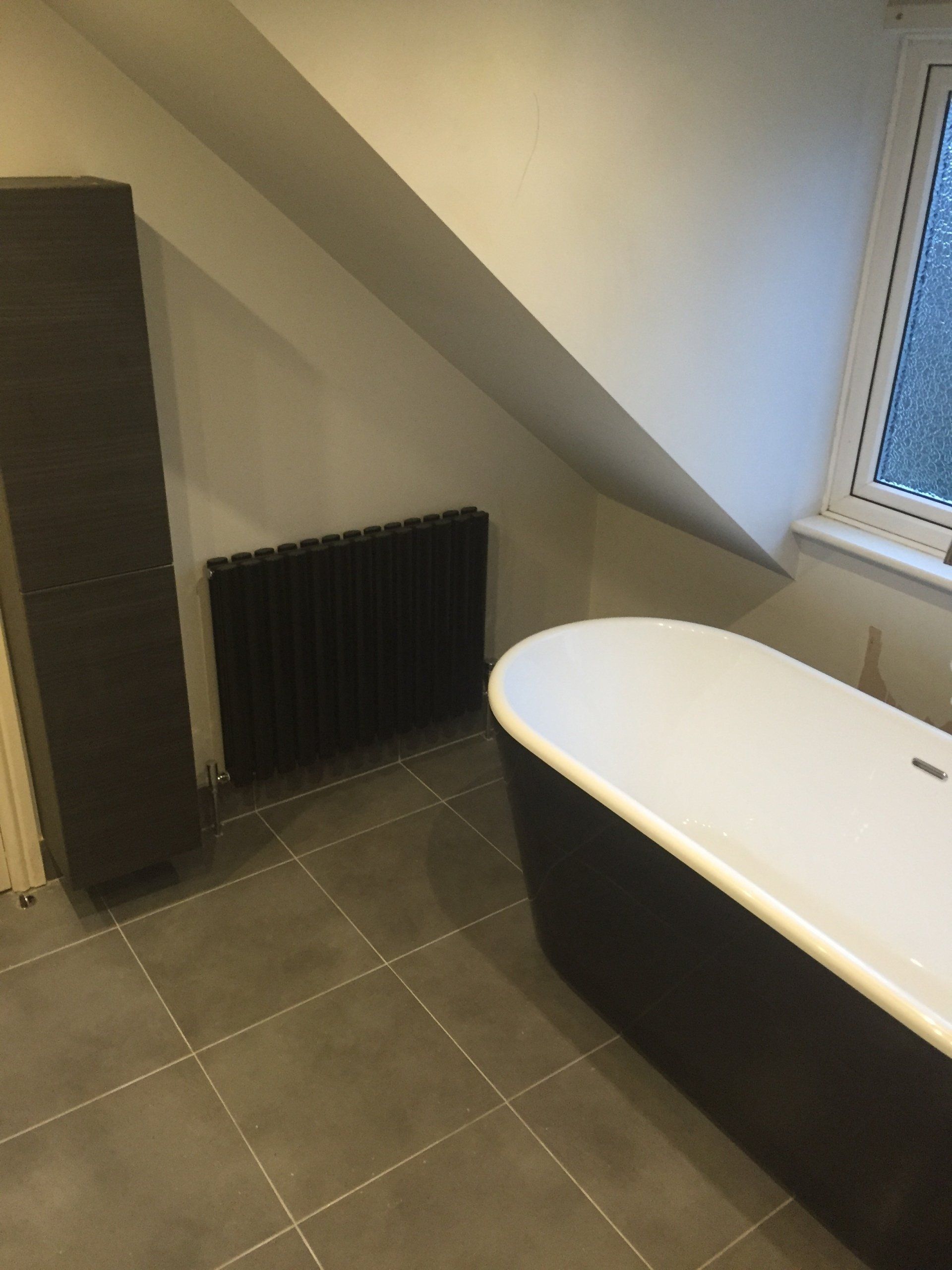Bathroom with black freestanding tub, dark radiator, and angled ceiling. Gray tiled floor.
