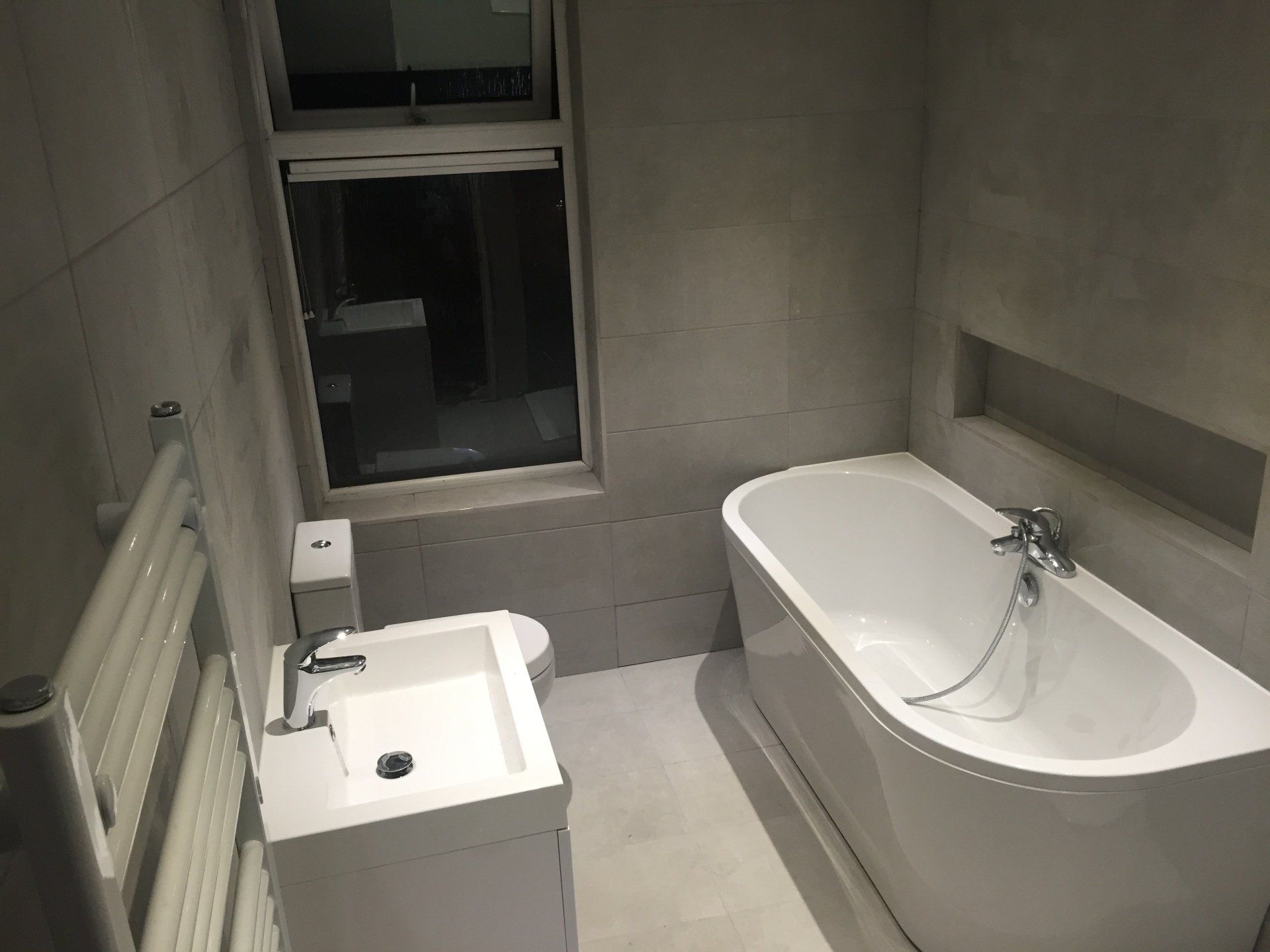 Bathroom with white tub, sink, and toilet, gray tiled walls. Window in the background.